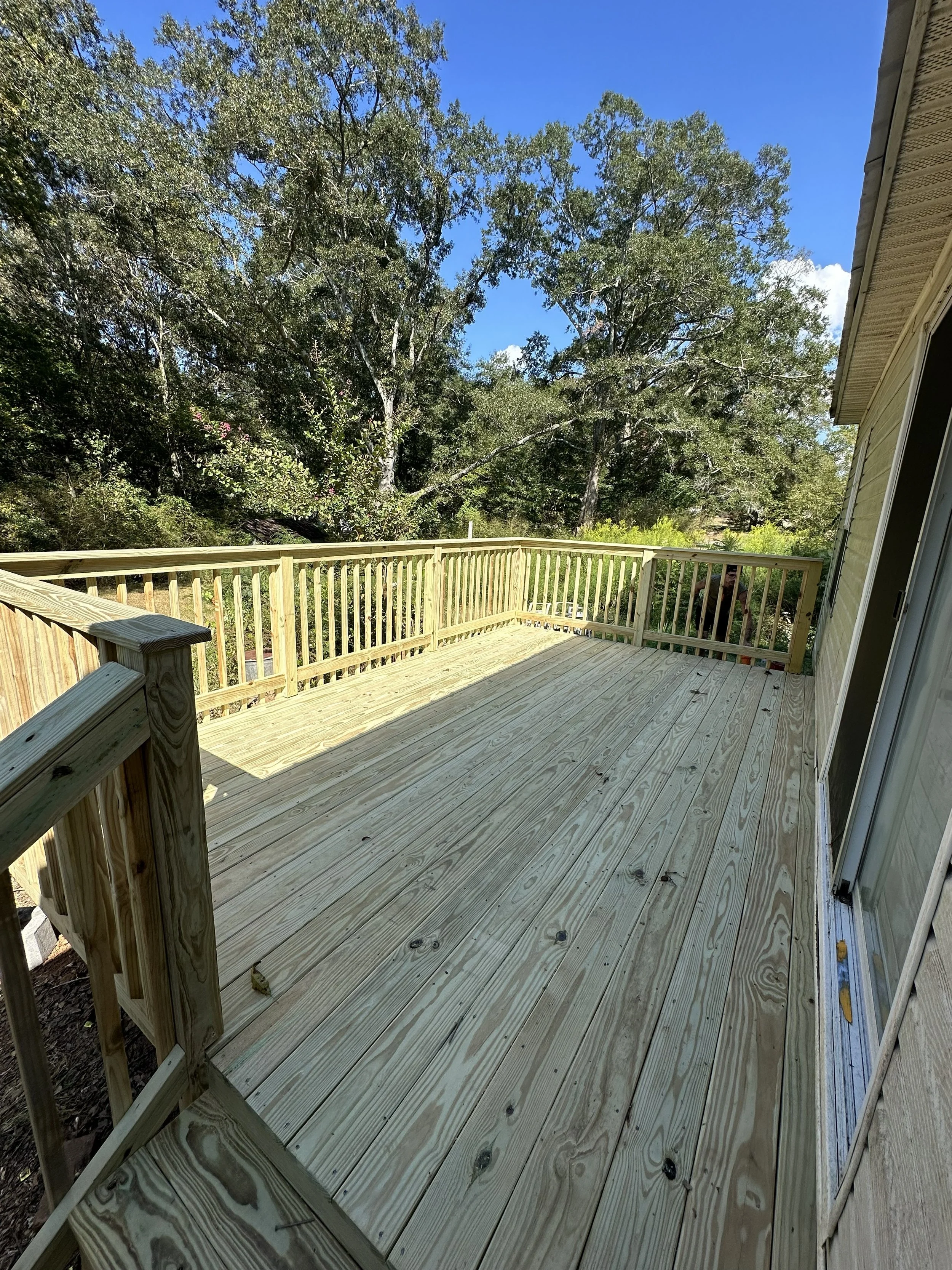 Newly constructed wooden deck attached to a house, surrounded by trees and a clear blue sky.