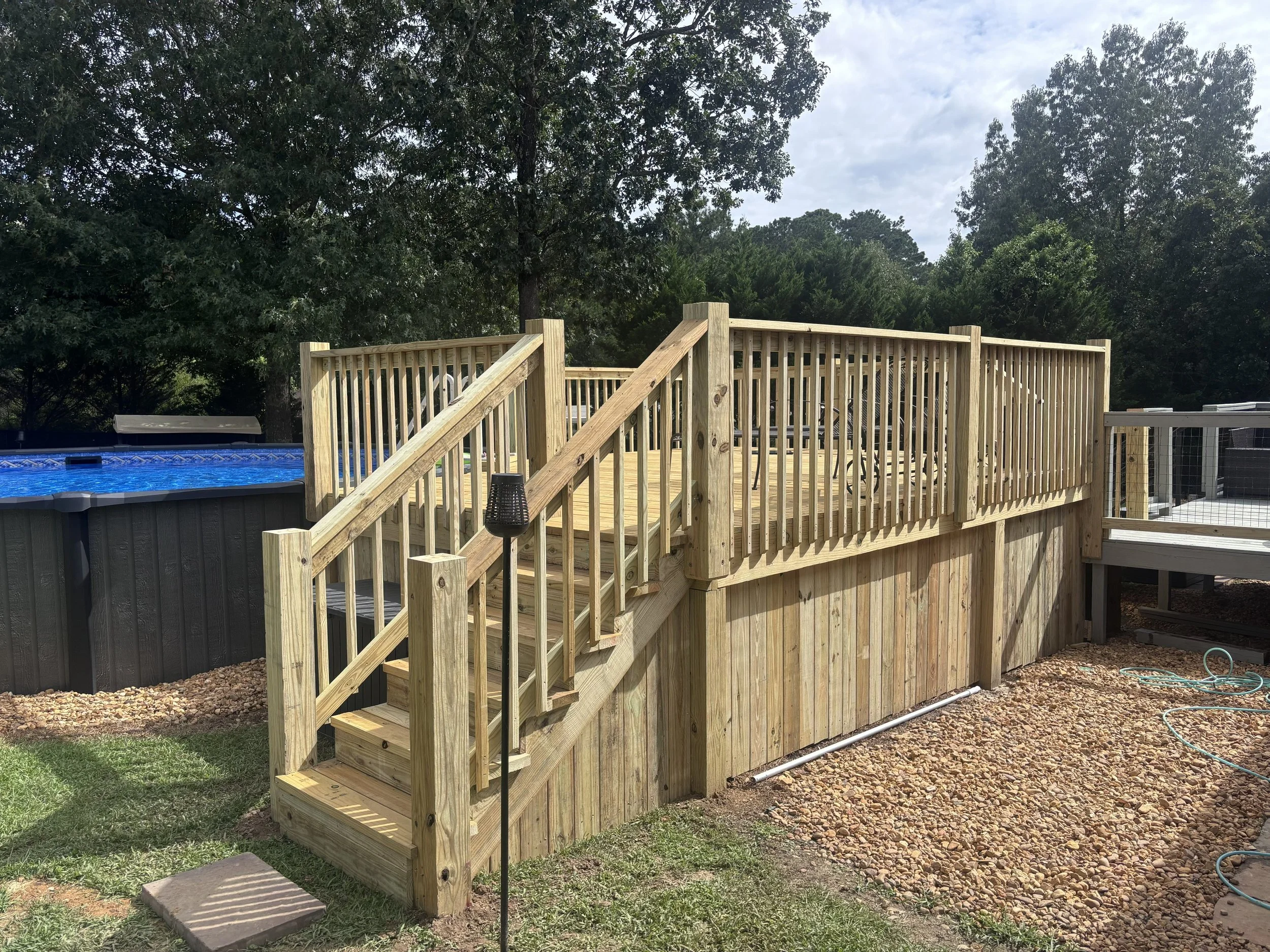 A wooden deck with stairs leading to a backyard, enclosed by a wooden railing, overlooking a tree-filled area under a partly cloudy sky.
