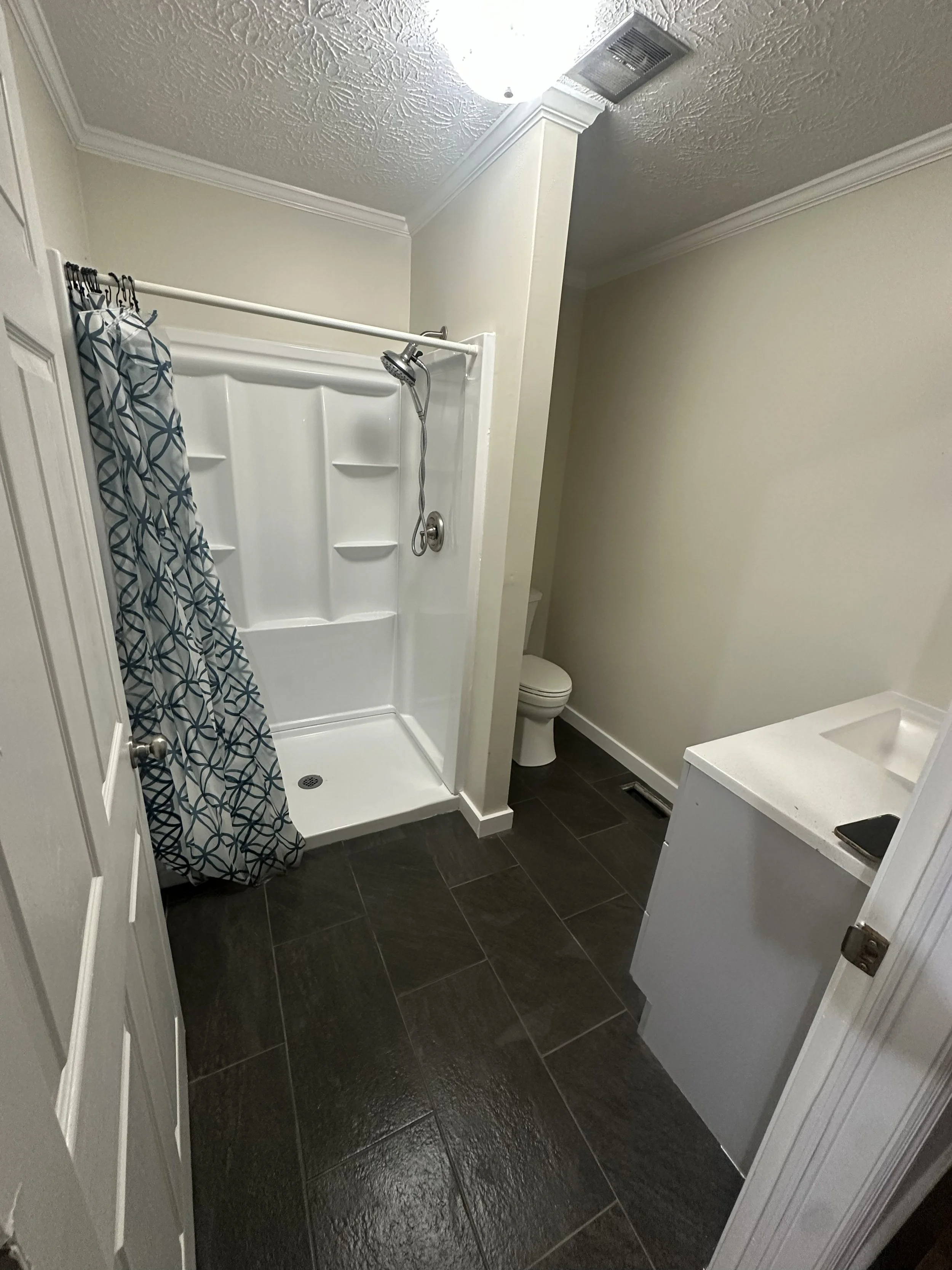 A small bathroom with a shower, toilet, and vanity with a sink. The shower has a patterned curtain, and the floor is tiled in dark color. The walls are painted light, and the ceiling is textured. There is a vent and light fixture on the ceiling.