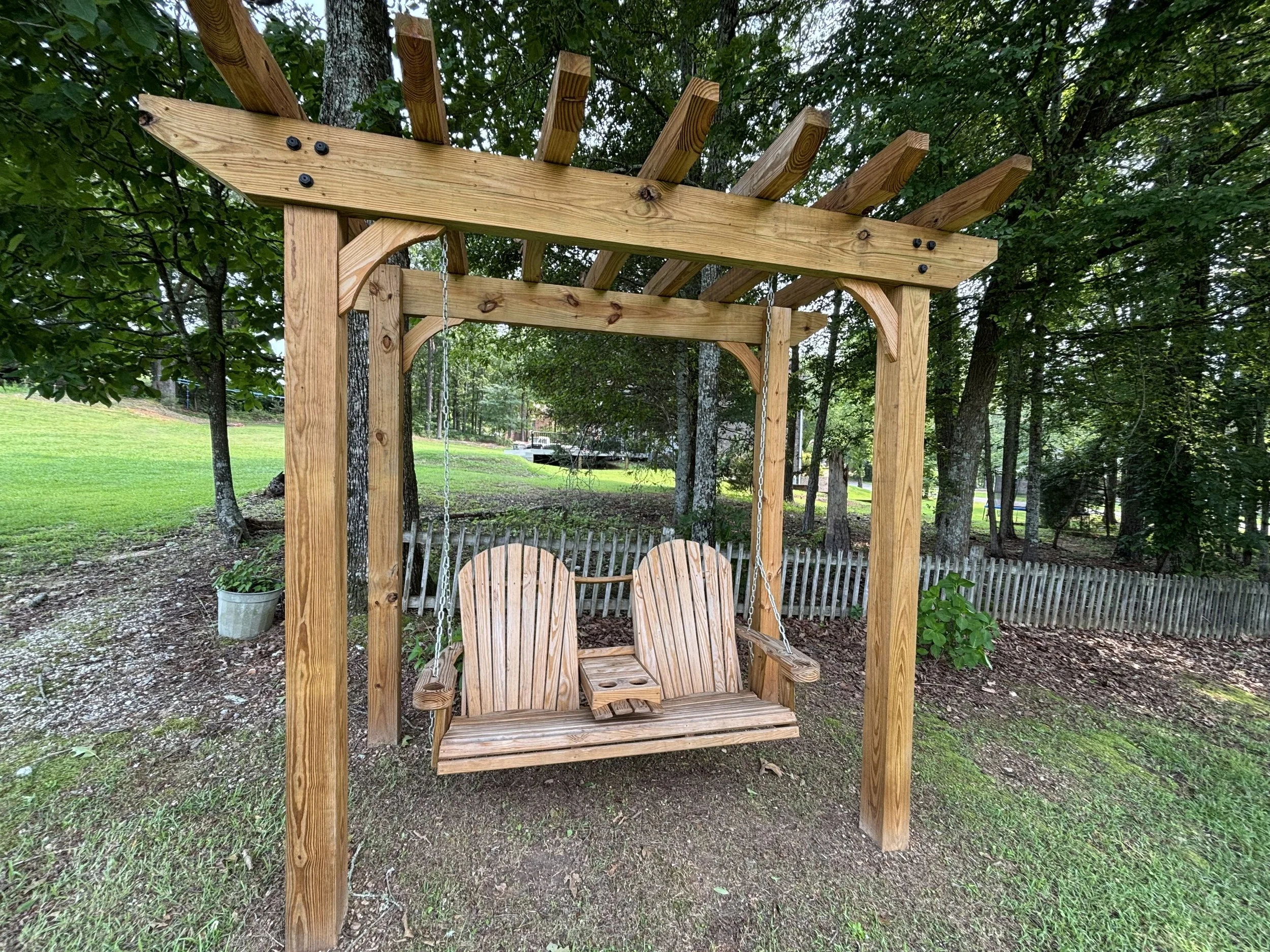 A wooden porch swing with a canopy frame, hanging from a wooden structure, set outdoors among trees and greenery.