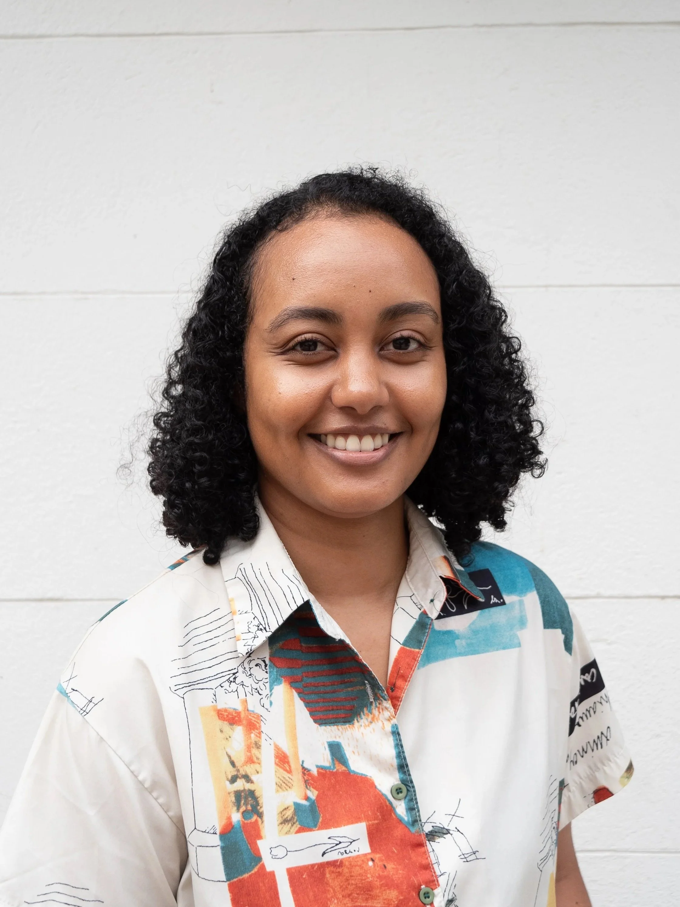 A smiling woman with curly black hair standing in front of a white wall, wearing a colorful patterned shirt with abstract designs.