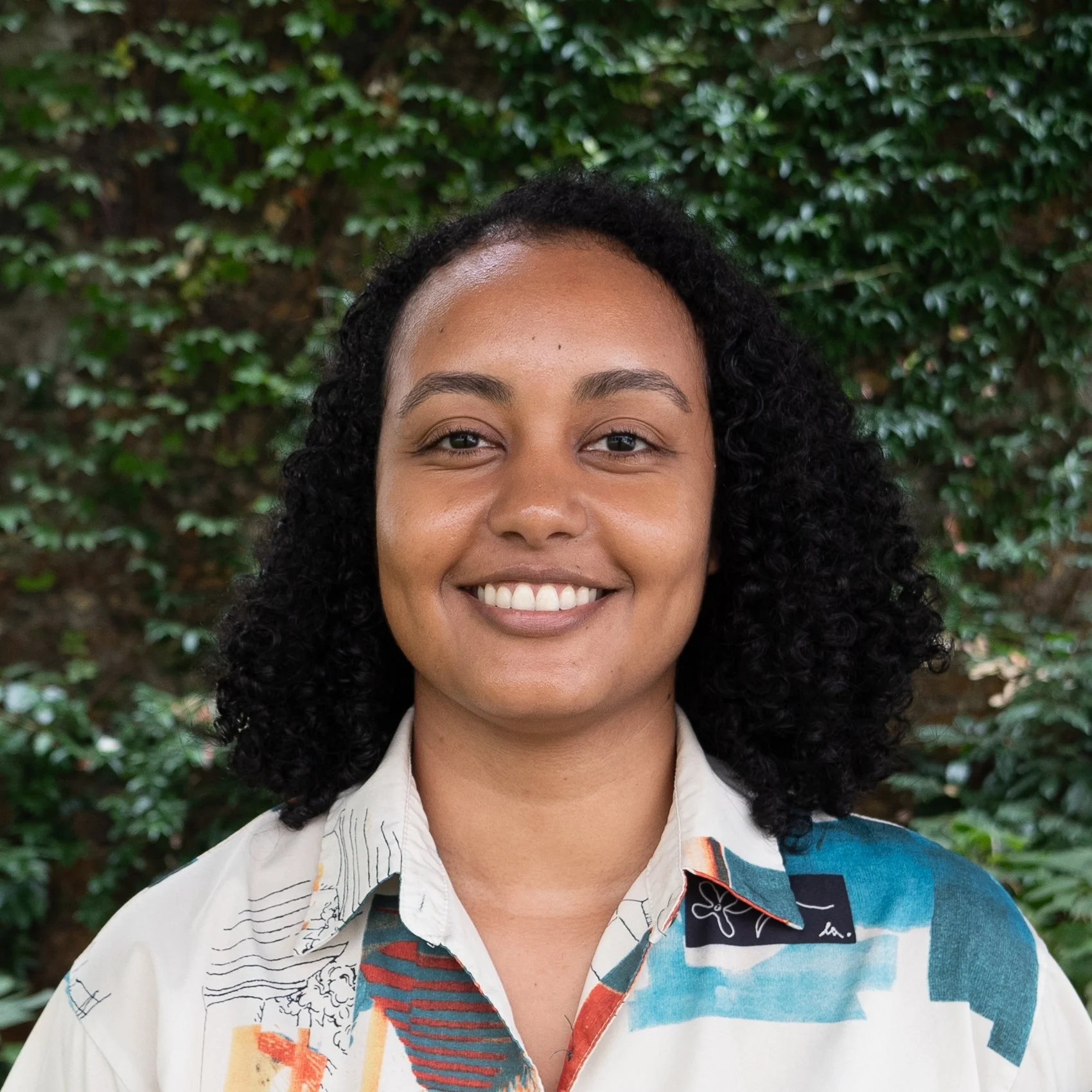 A woman with curly black hair smiling in front of a leafy green background.