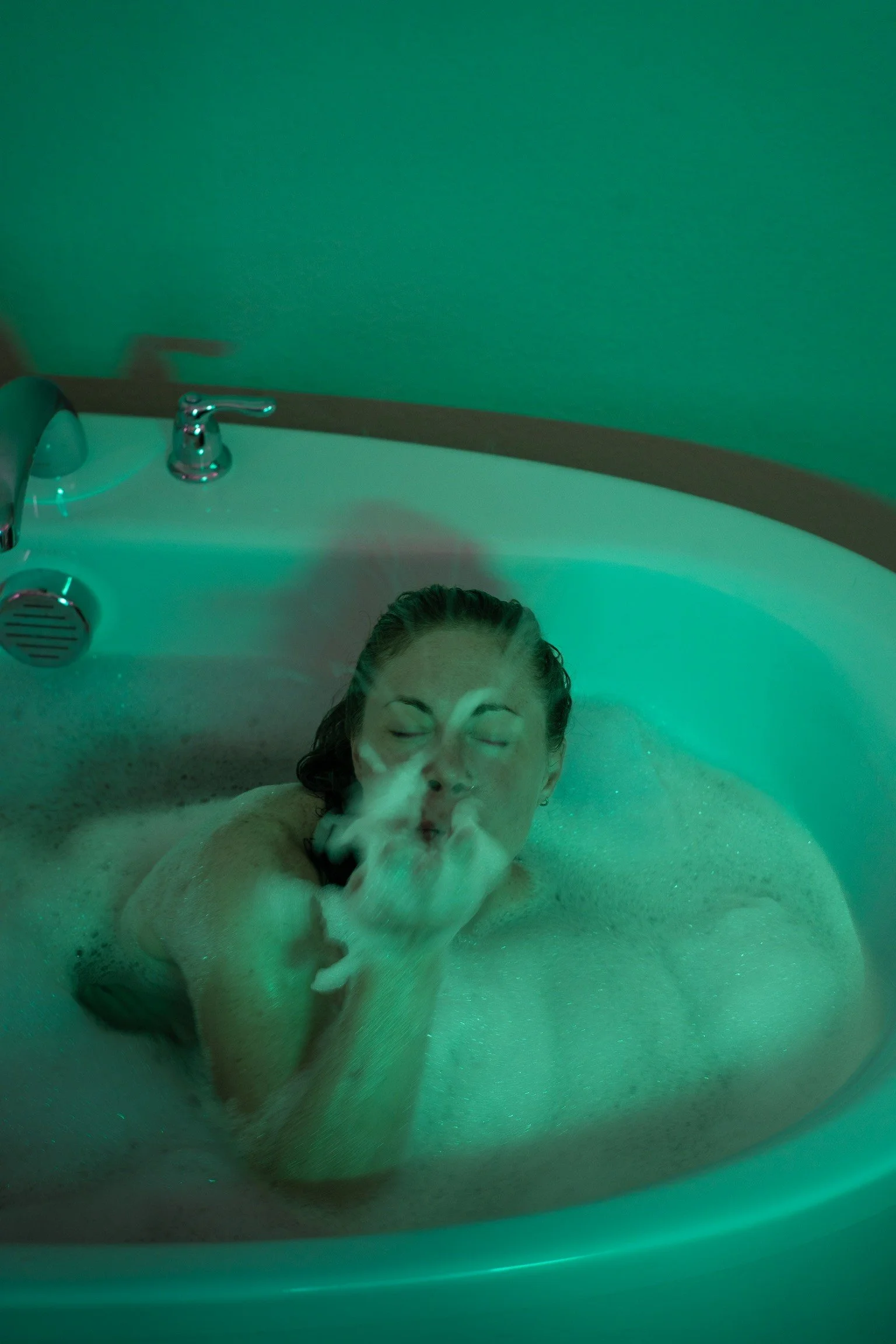 A person relaxing in a bubble bath with green lighting, with only the top of their head and face visible, eyes closed, and hair wet.