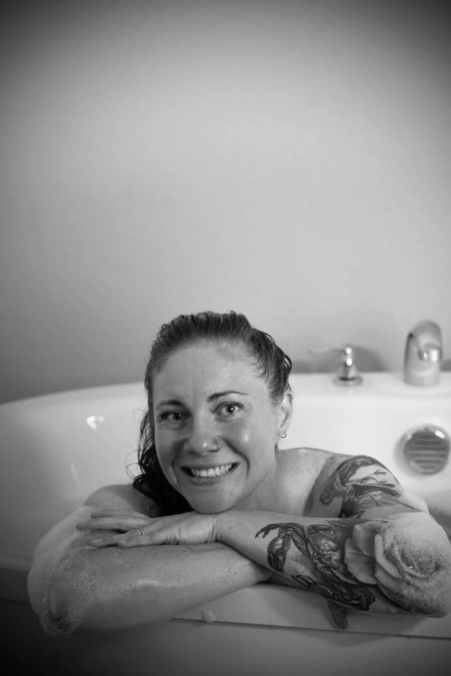 A woman with tattoos smiling while leaning on the edge of a bubble bath or hot tub in a bathroom with a sink and faucet in the background.