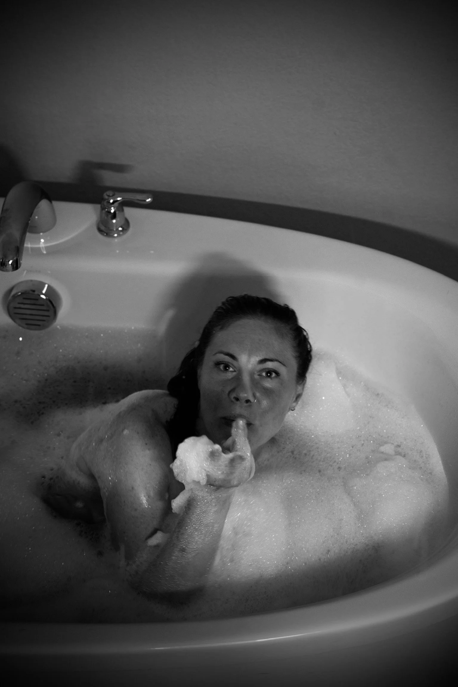 A woman with wet hair inside a bathtub filled with bubbles, holding a foam object, with a sink faucet and drain visible in the background.