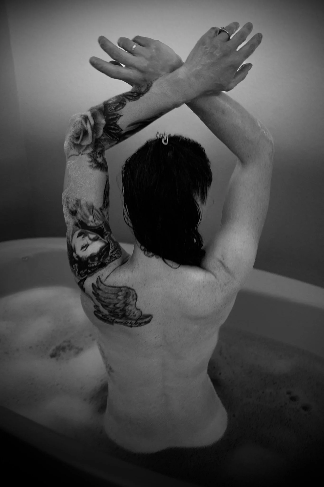 A person with tattoos on their arm, with dark hair, is sitting in a bathtub, with arms crossed behind their head.