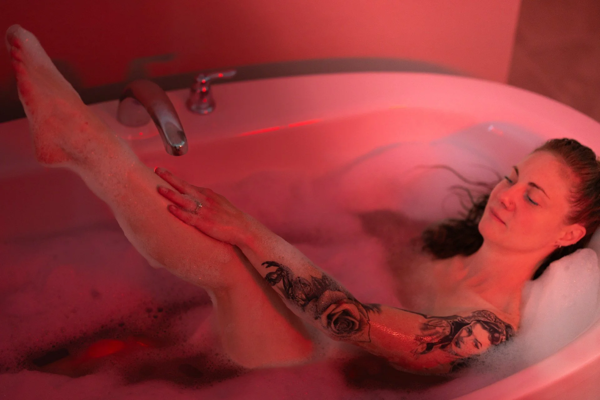 A woman relaxing in a bubble bath with red lighting, showing her smiling face and tattooed arm resting on the edge of the tub.