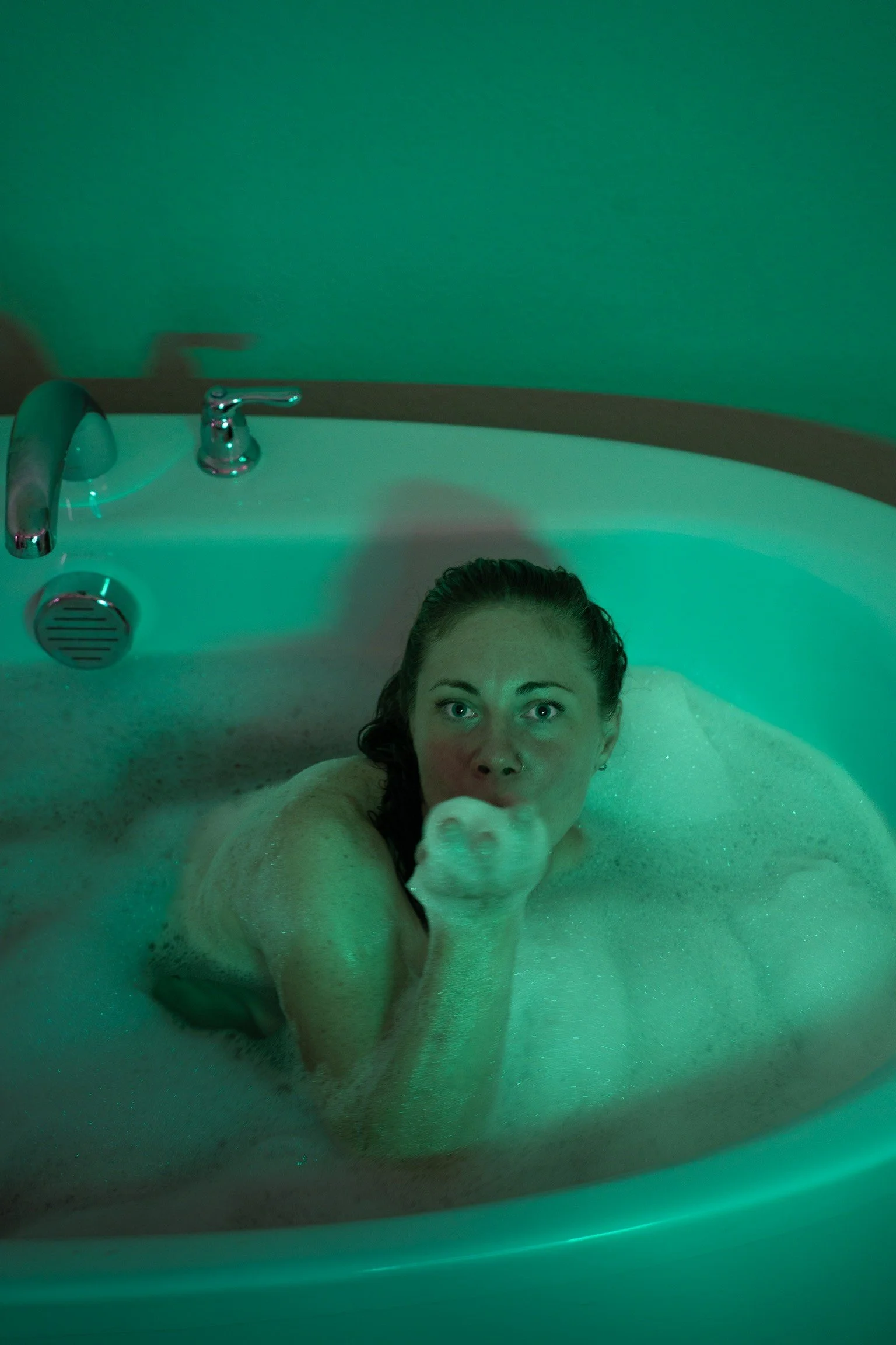 A woman with wet hair taking a selfie while relaxing in a bubble-filled bathtub illuminated by green lighting.