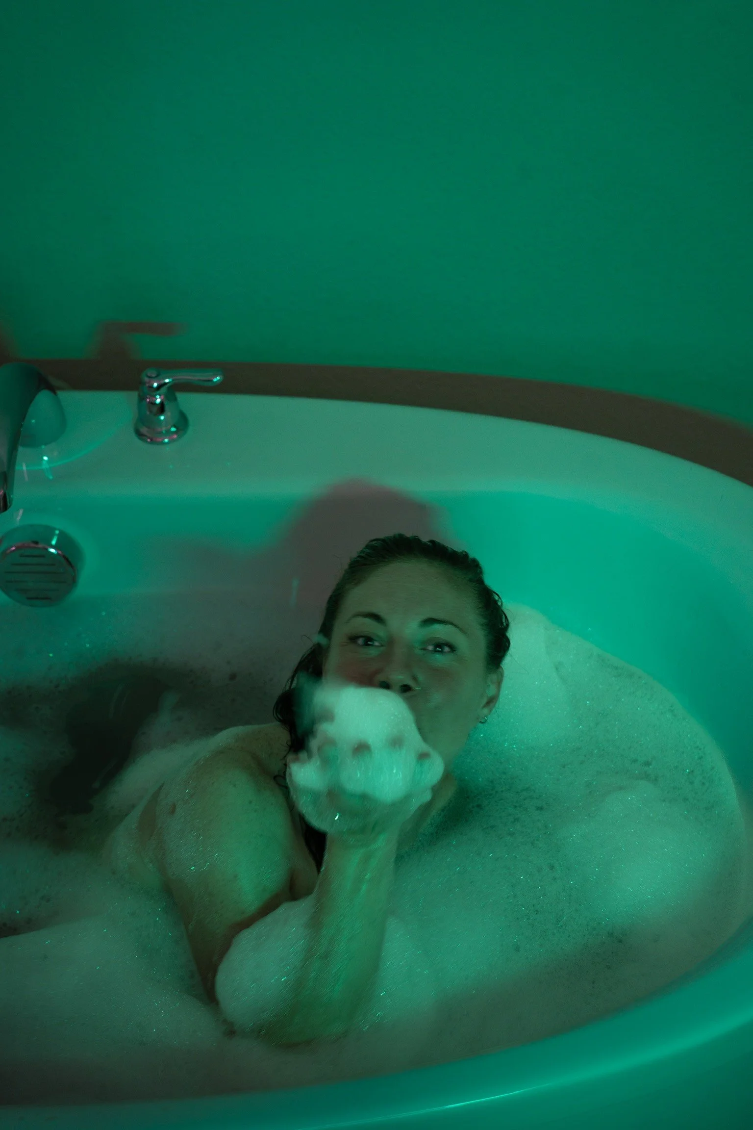 A woman with wet hair relaxing in a bubble bath with a dog, surrounded by green lighting.