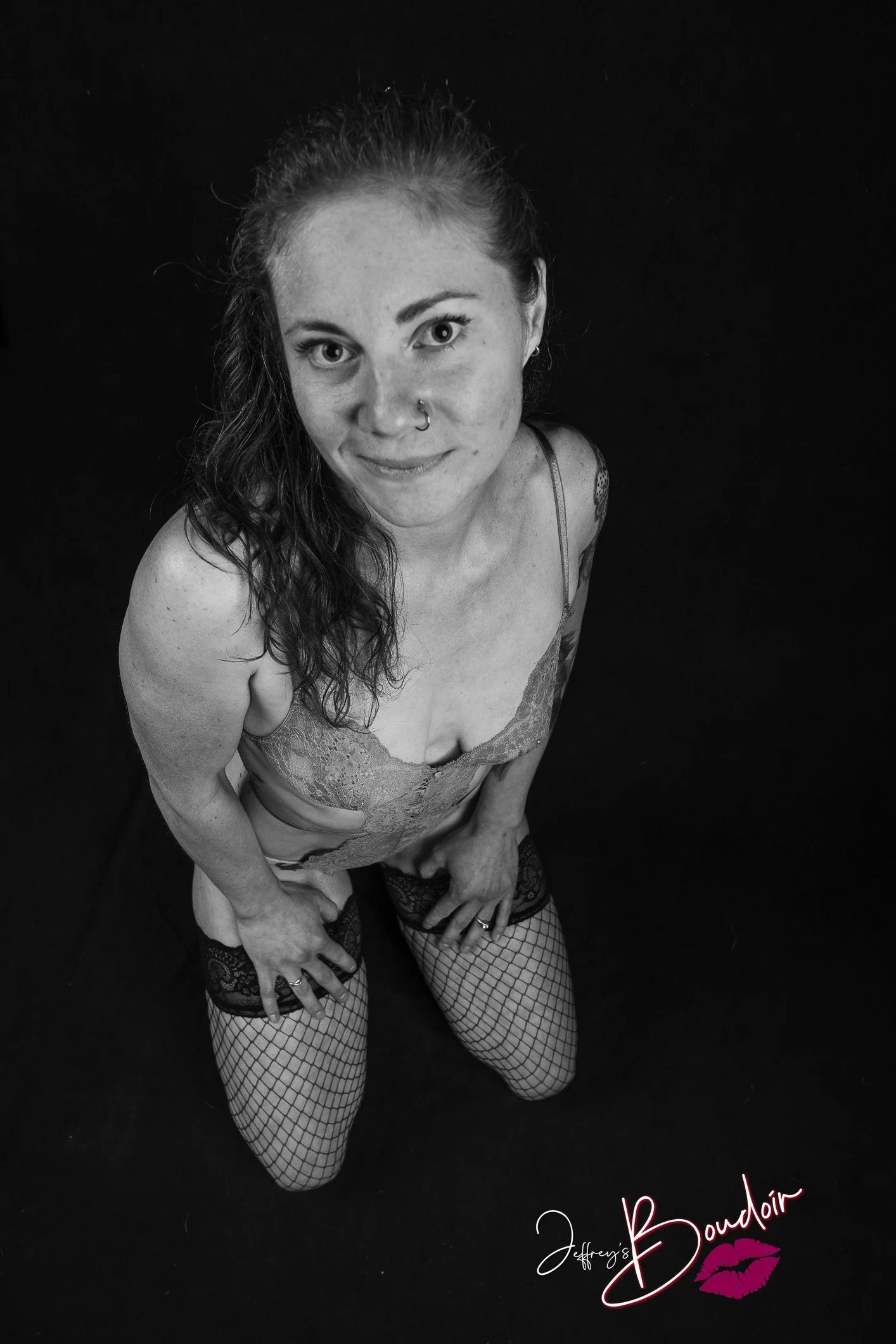 A woman kneeling on a dark surface, looking up at the camera, wearing lingerie and fishnet stockings, with text reading 'Jeffrey's Boudoir' and a kiss mark at the bottom.