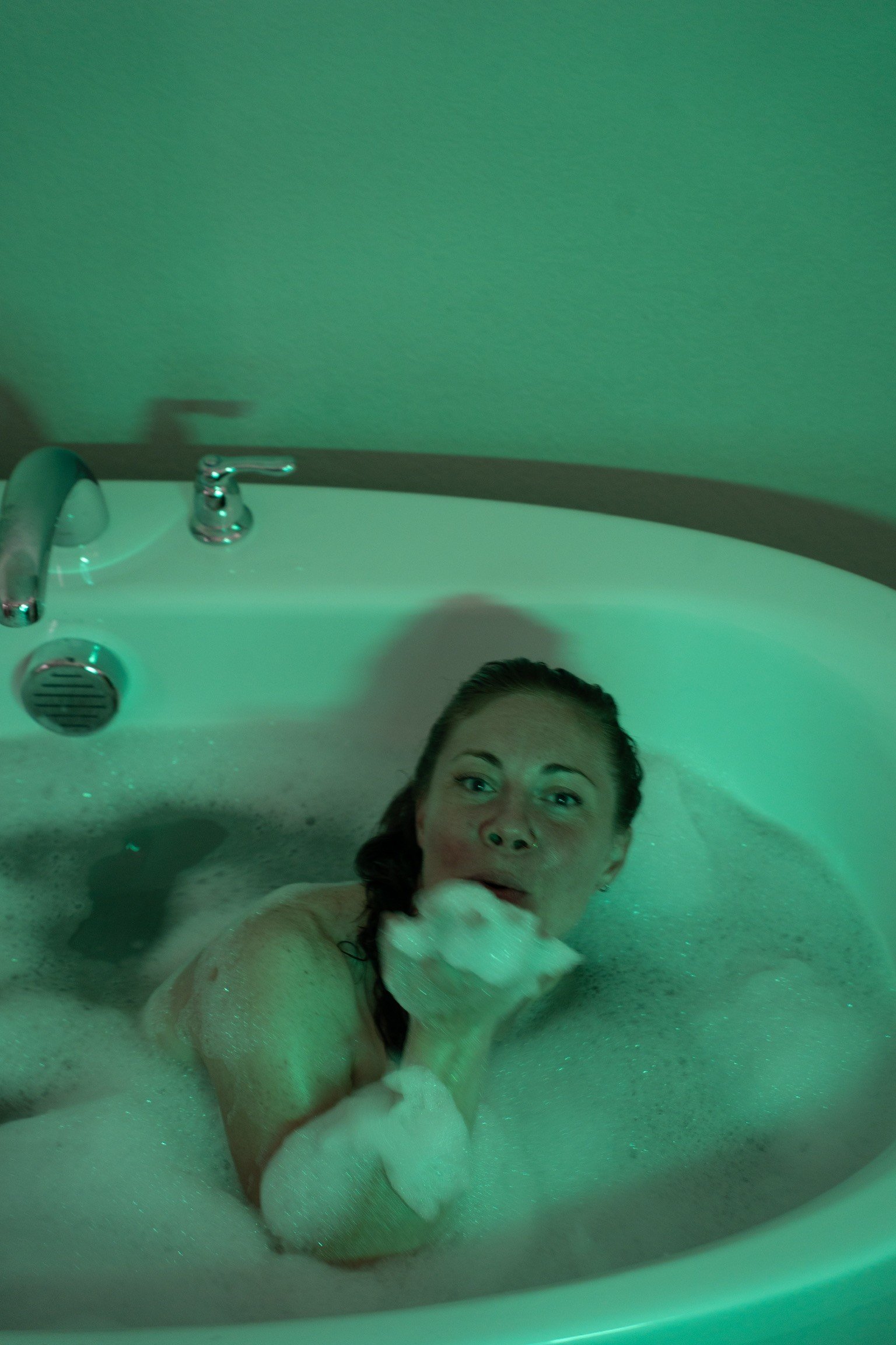 Woman taking a bubble bath in a white bathtub with green lighting.