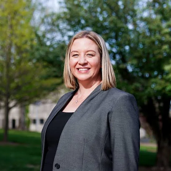 A smiling woman with shoulder-length blonde hair wearing a gray blazer standing outdoors in front of green trees.
