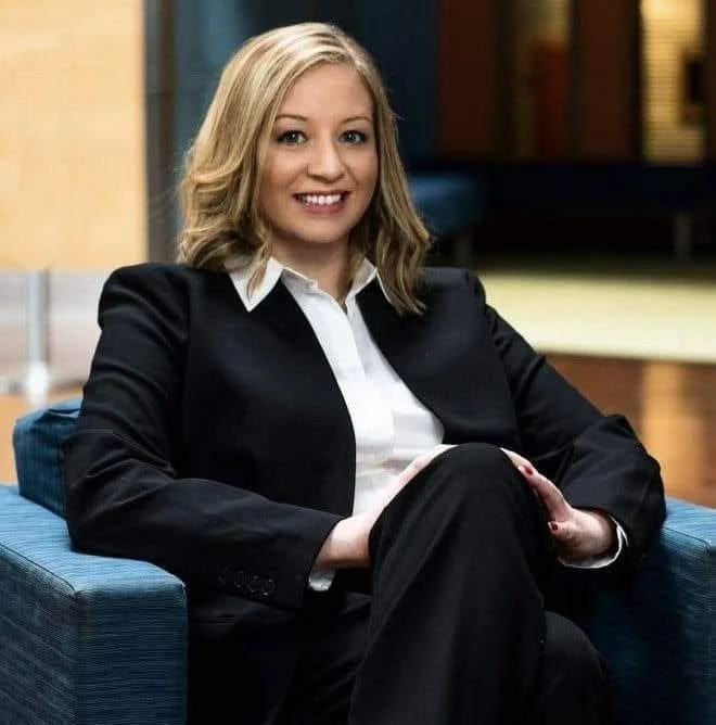 A woman with blonde hair smiling while sitting on a blue armchair in a professional setting, wearing a black blazer over a white shirt.