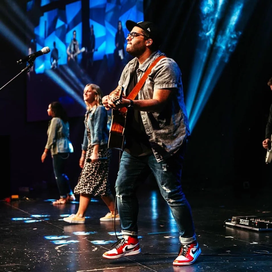 Musician playing guitar on stage with backup singers and band members, colorful stage lighting