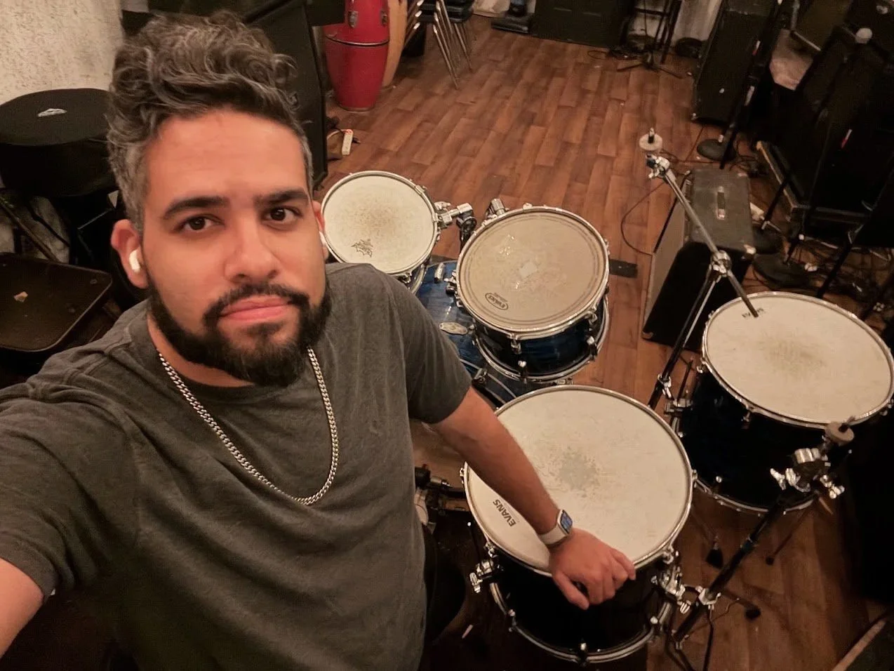 A man with dark hair and a beard, wearing headphones, a gray t-shirt, and a chain necklace, is taking a selfie in a music studio, standing behind a drum set with multiple drums and cymbals, surrounded by various music equipment and wooden flooring.
