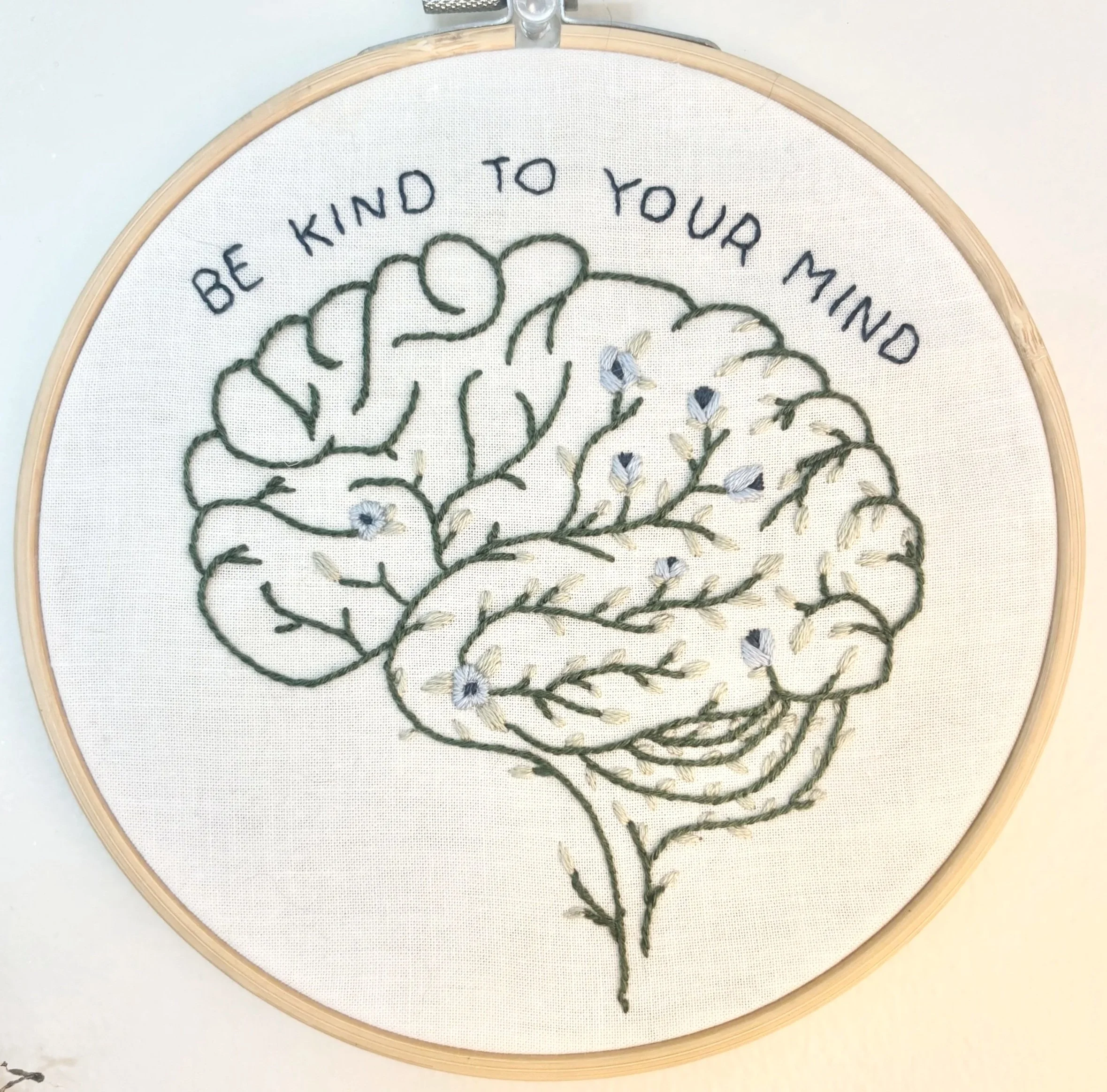 Embroidery hoop with a design of a brain and floral elements above it. Text embroidered around the top reads, 'BE KIND TO YOUR MIND'.