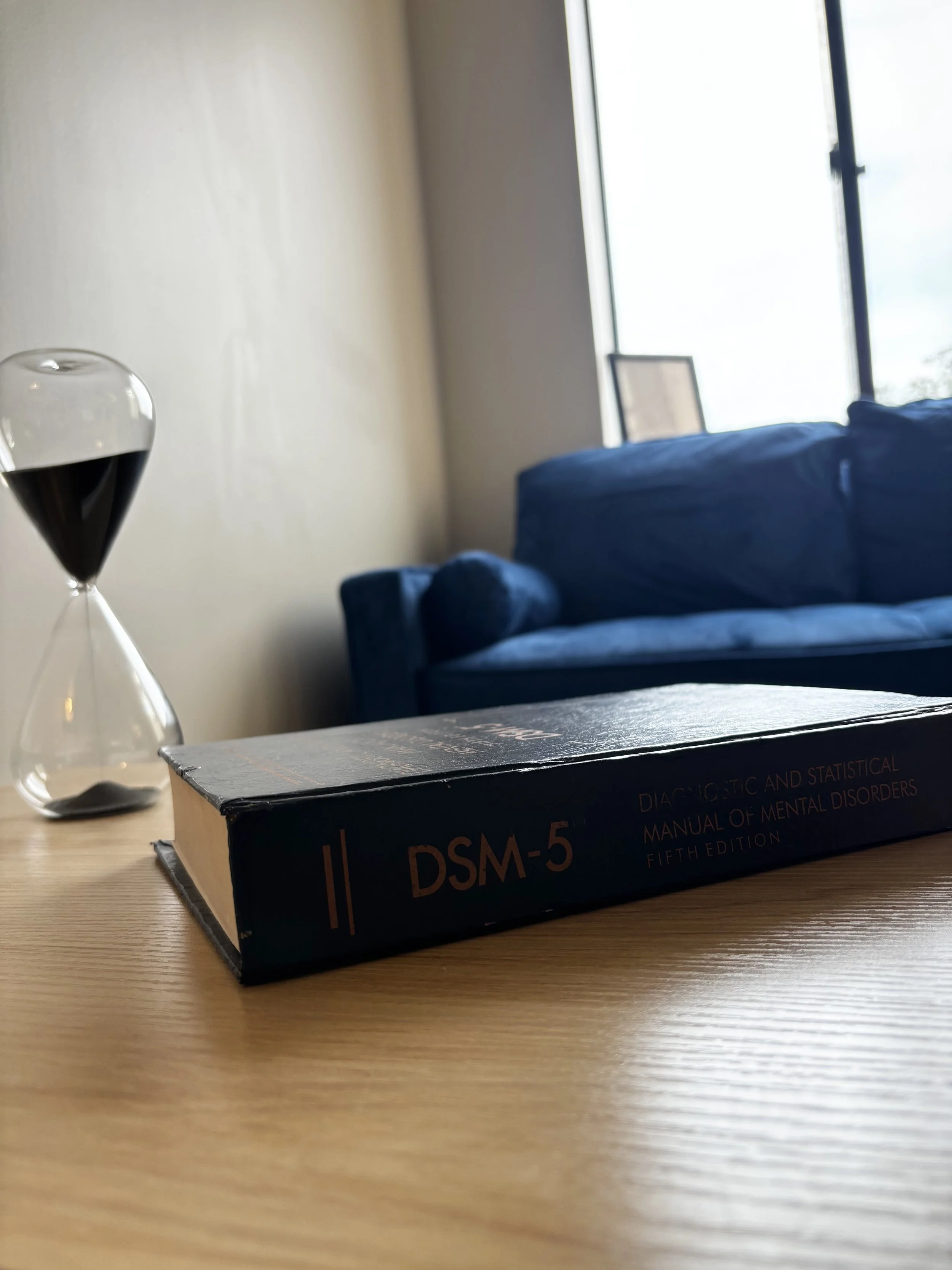 Book titled 'DSM-5 Diagnostic and Statistical Manual of Mental Disorders, Fifth Edition' on a wooden table, with a glass of dark-colored beverage and a blurred blue sofa and window in the background.
