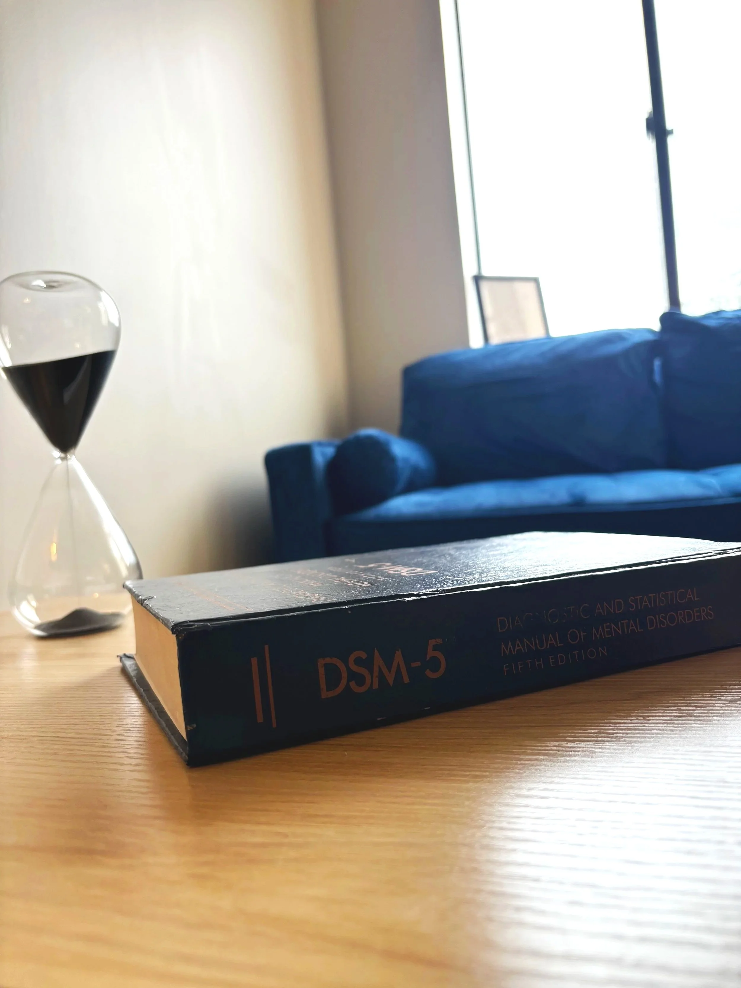 A blue book titled "DSM-5" on a wooden table, with a glass of dark liquid and a blurred background including a blue couch and a window.