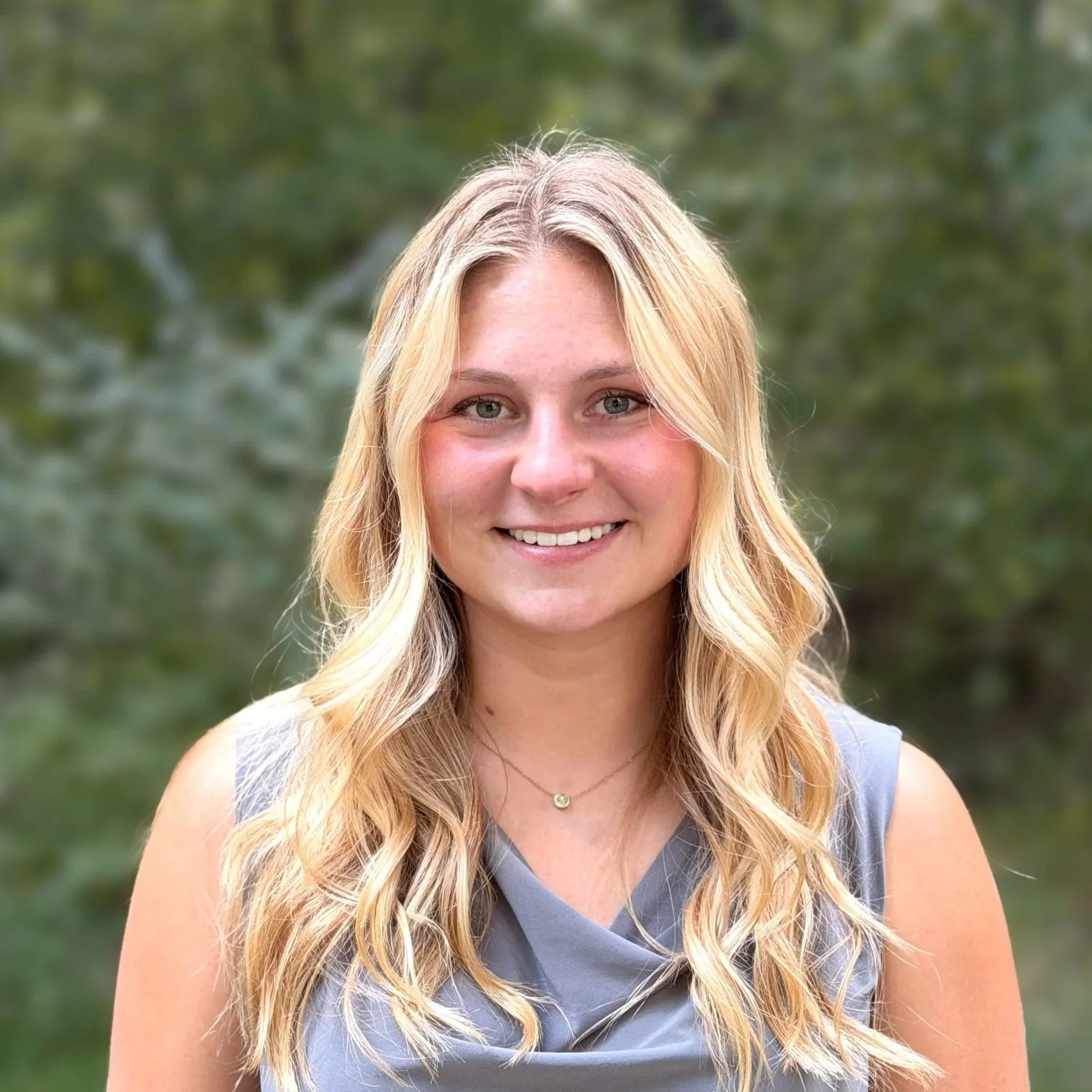 Close-up of a smiling woman with long blonde wavy hair, wearing a silver necklace and sleeveless top, standing outdoors with blurred green trees in the background.
