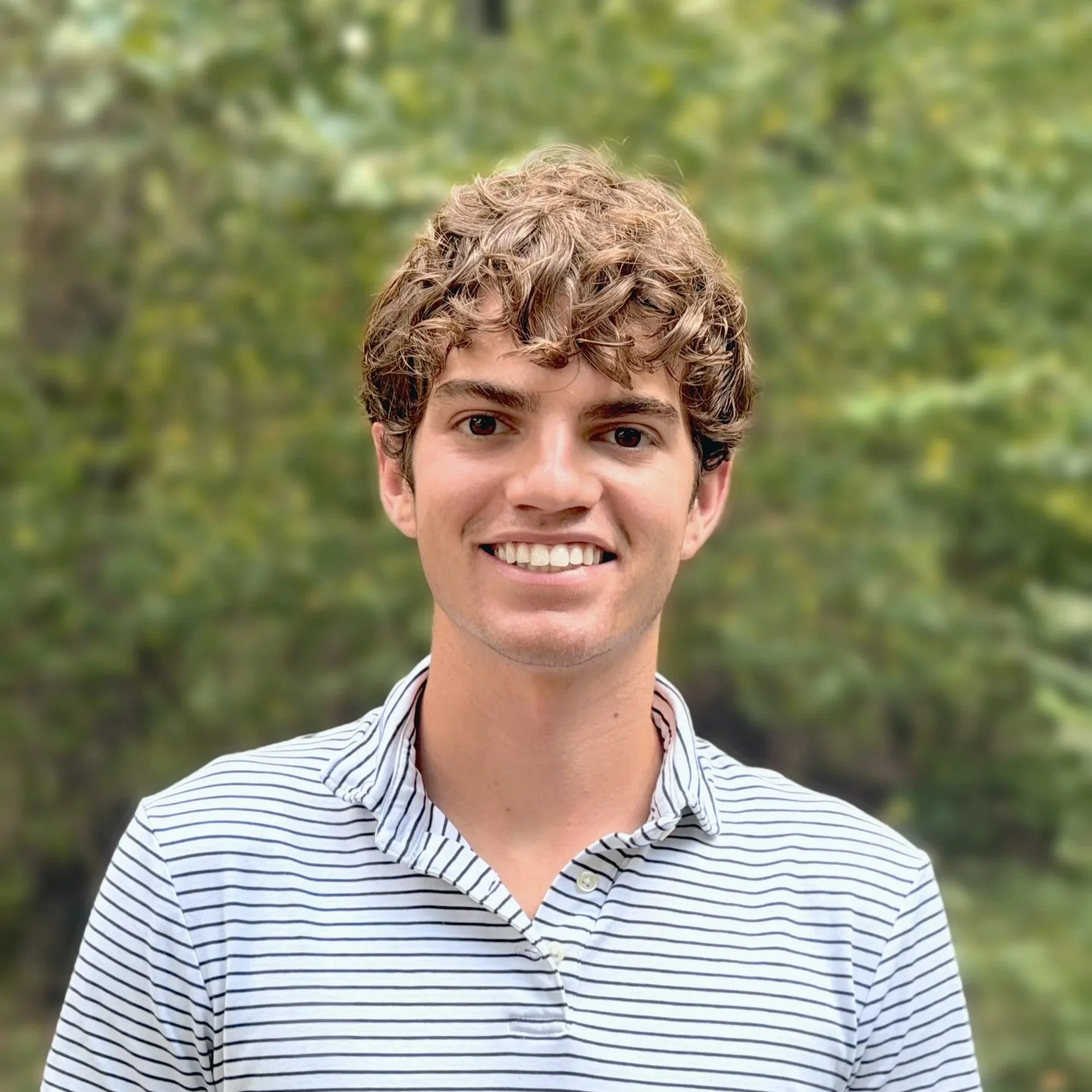Close-up of a young man's face with curly hair, smiling, wearing a striped shirt outdoors with green trees in the background.