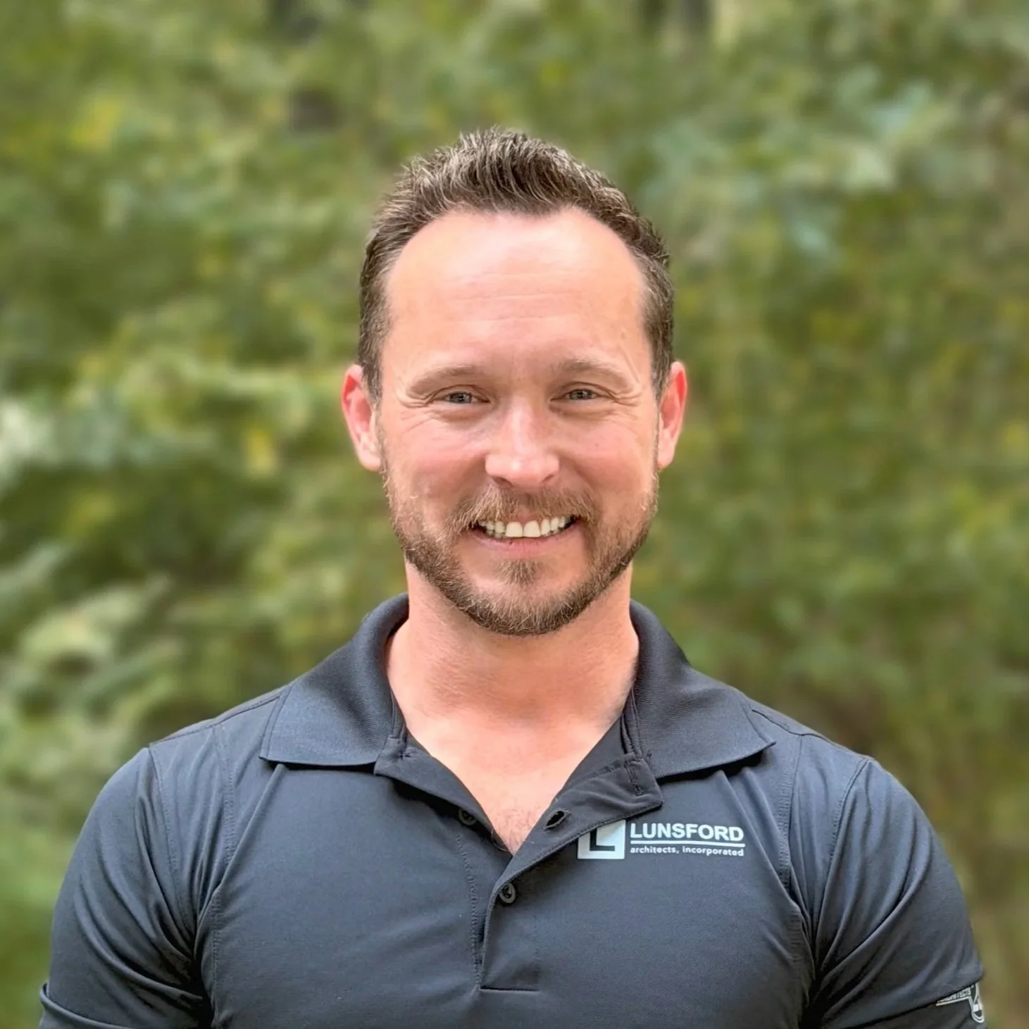 A smiling man with a beard and short dark hair, wearing a black collared shirt with a logo and text that reads 'LUNSFORD architects, incorporated,' standing outdoors with blurred green trees in the background.