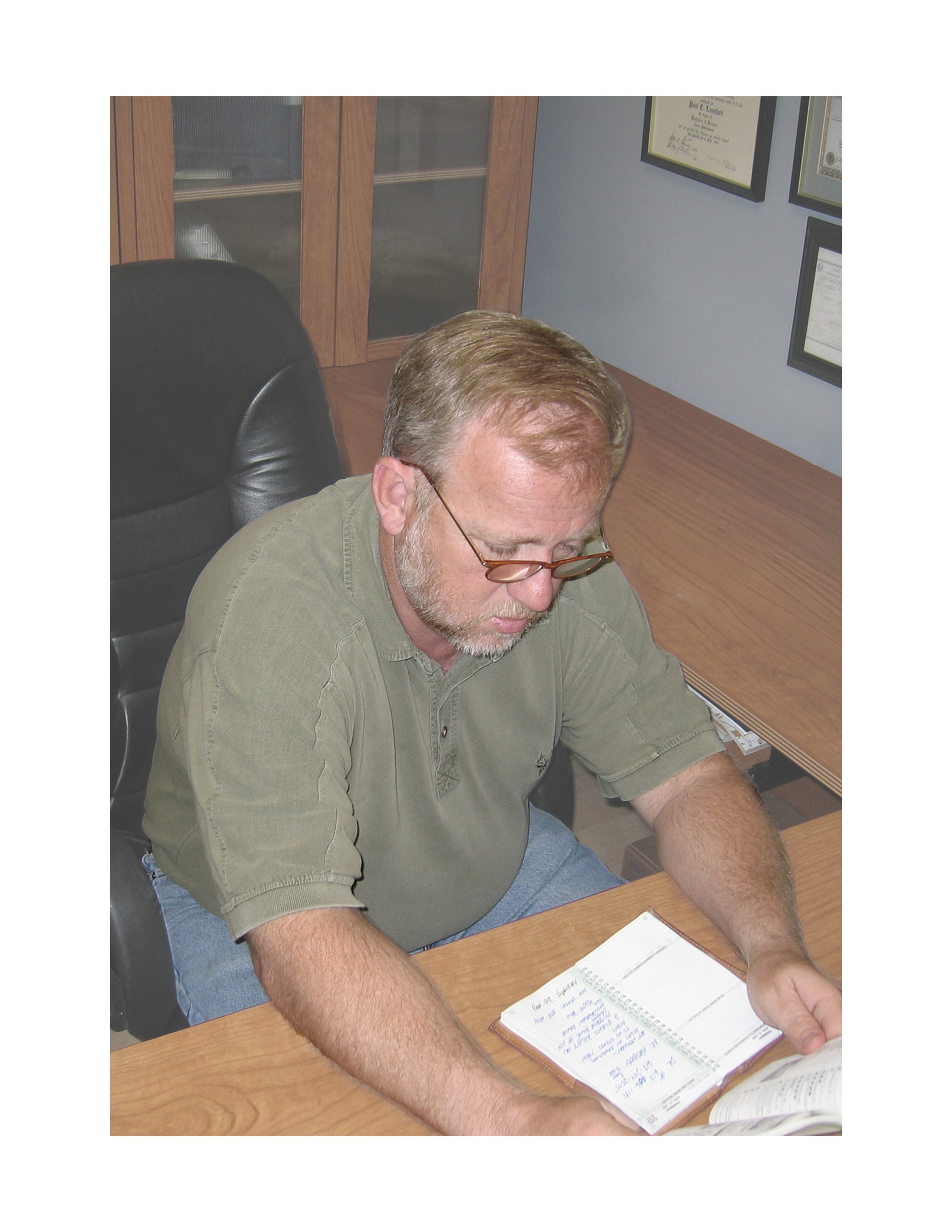 A man with glasses and a beard, sitting at a desk, reading a notebook with handwritten notes.
