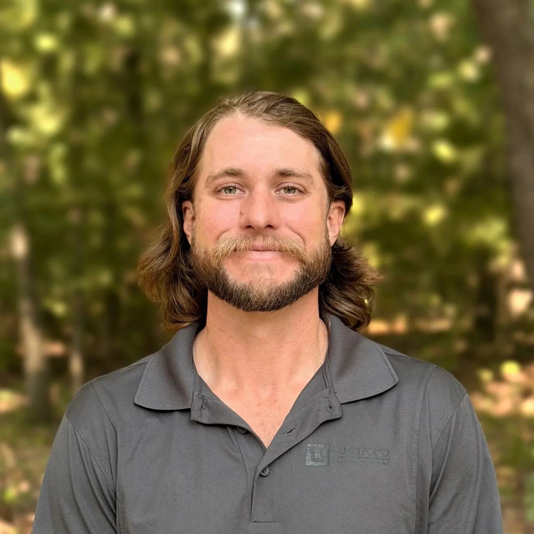 A man with long brown hair, a beard, and blue eyes smiling at the camera, wearing a dark gray polo shirt, outdoors with blurred green trees in the background.