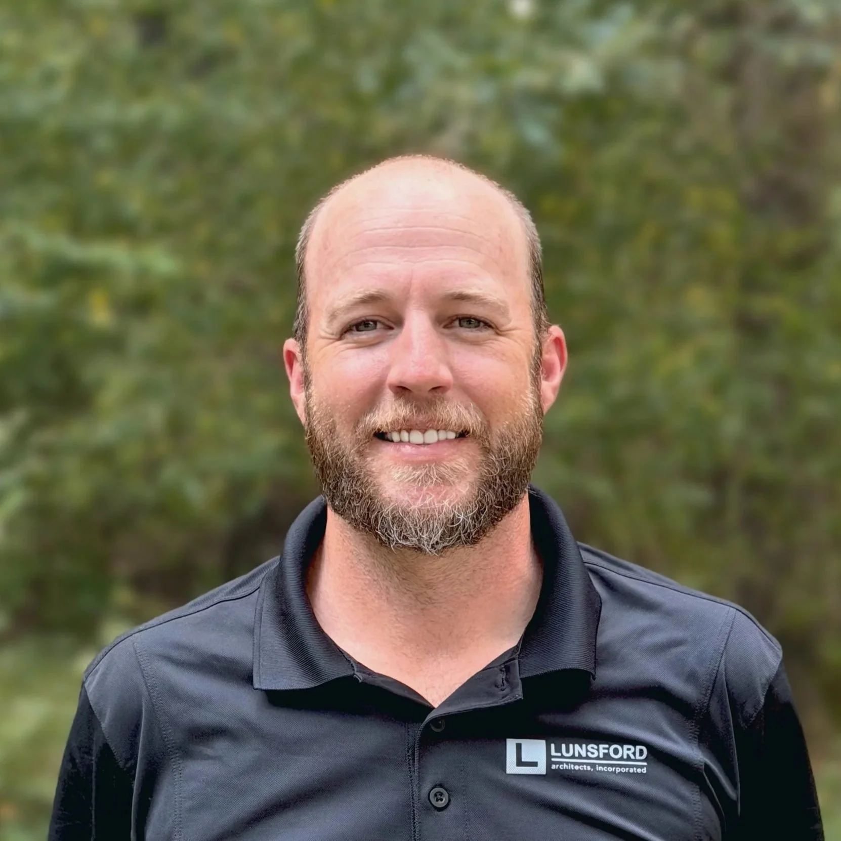 A smiling man with a beard and short hair, wearing a black collared shirt with a logo for Lunsford Architects, standing outdoors with blurred greenery in the background.