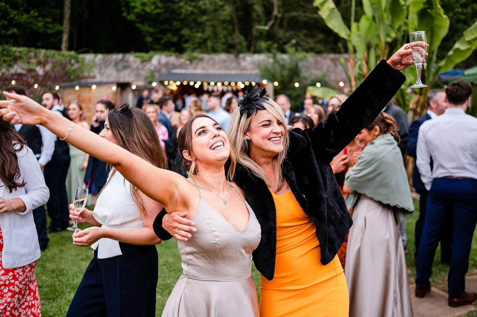 Two women smiling and celebrating at an outdoor gathering, holding glasses of champagne, surrounded by other guests in a lush garden setting.