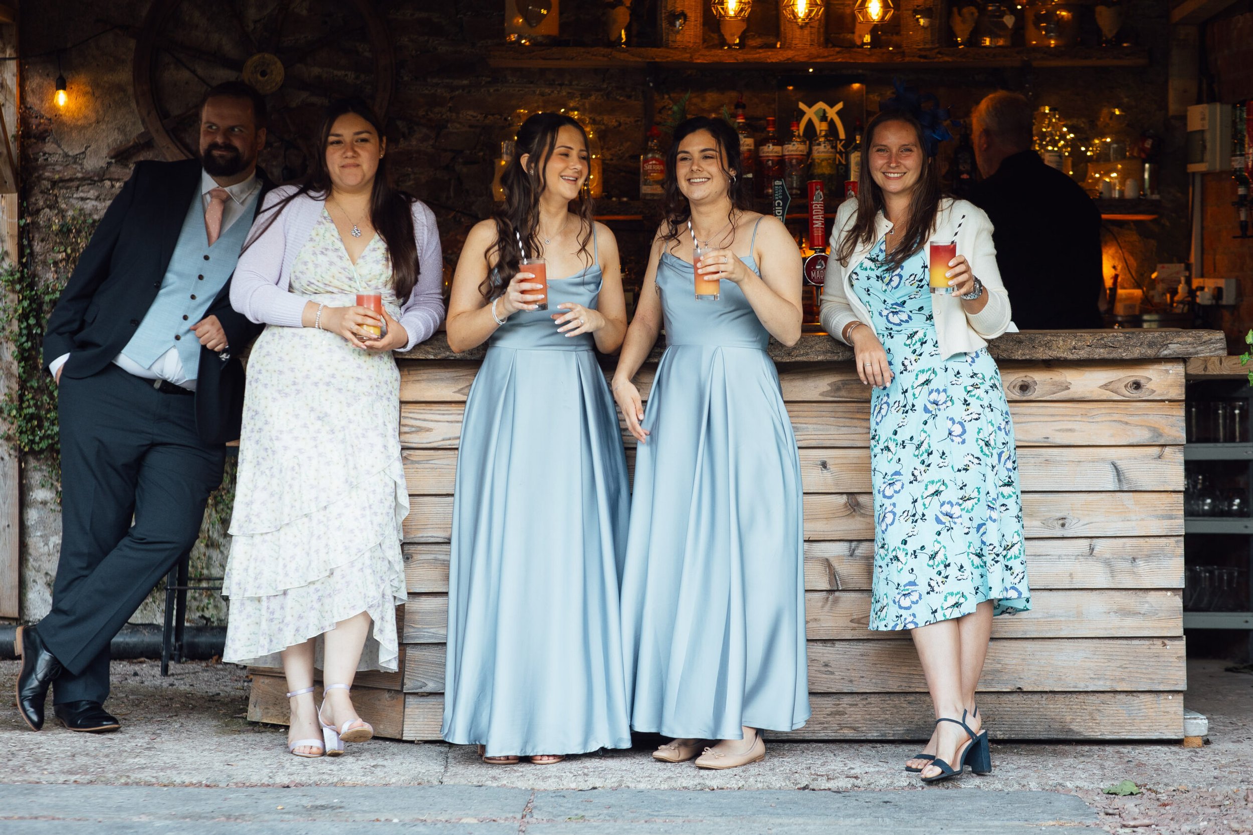 Group of six friends enjoying drinks at a bar at night, dressed in formal and semi-formal attire, smiling and socializing.