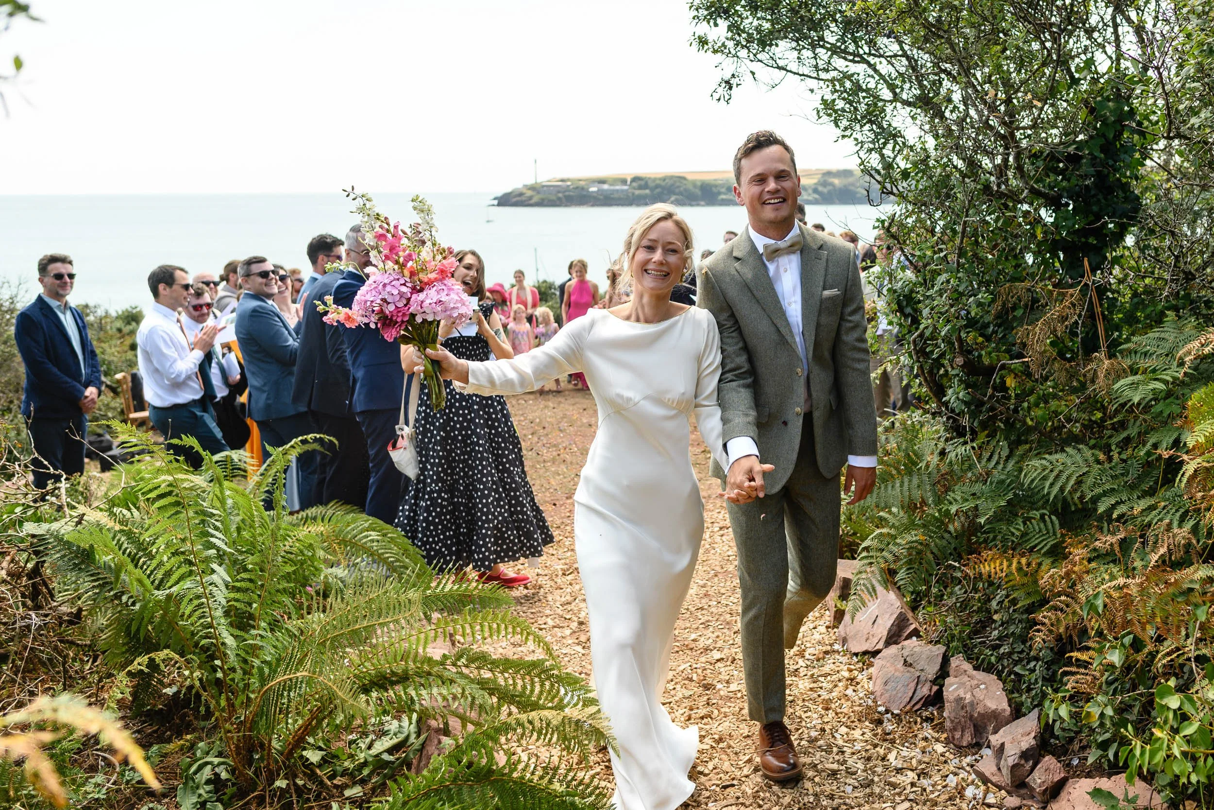 A bride and groom holding hands and walking outdoors at a wedding ceremony, with guests clapping in the background near a body of water and green landscape.
