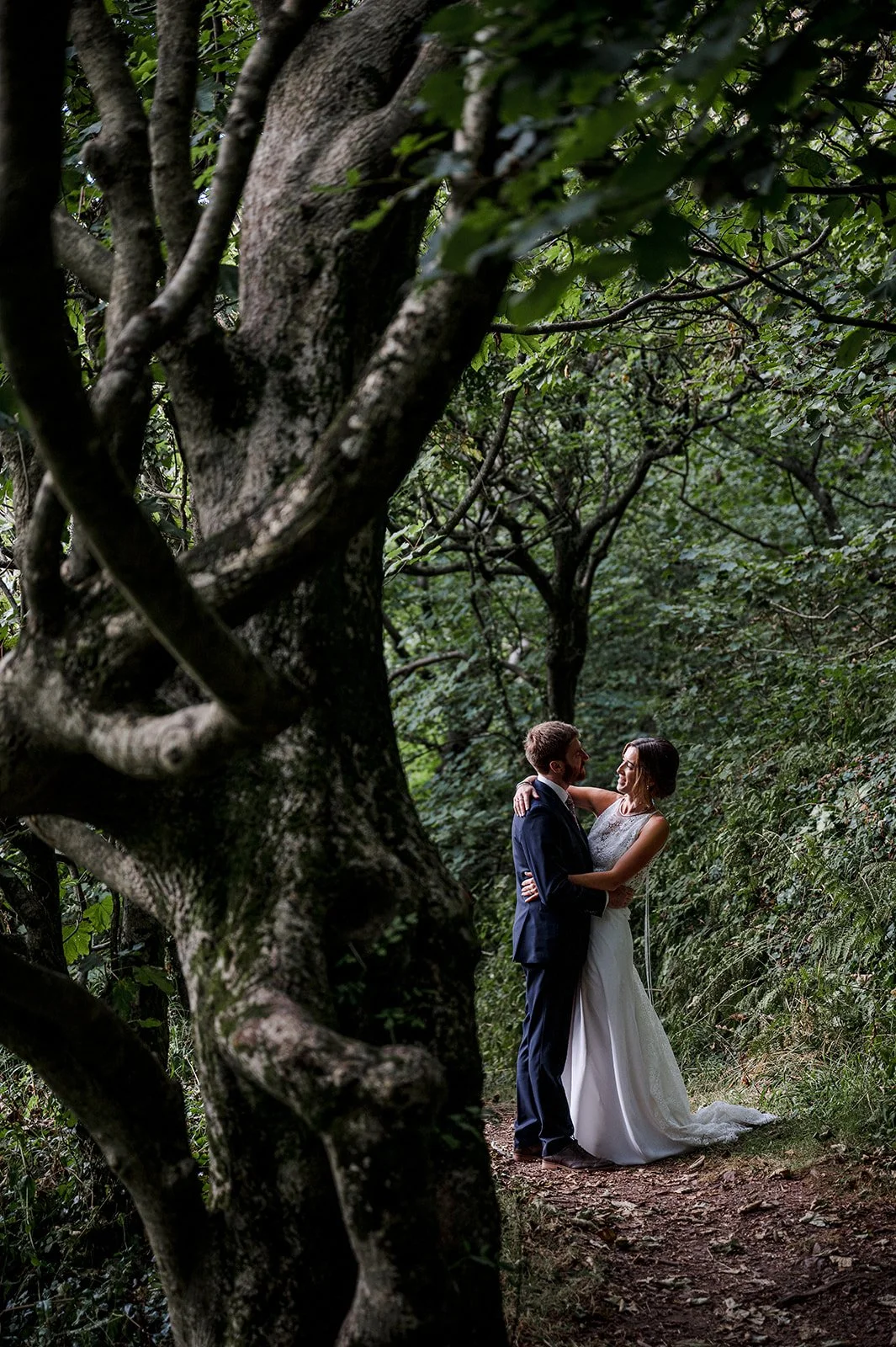 A bride and groom standing close together in a forest, embracing and gazing into each other's eyes.