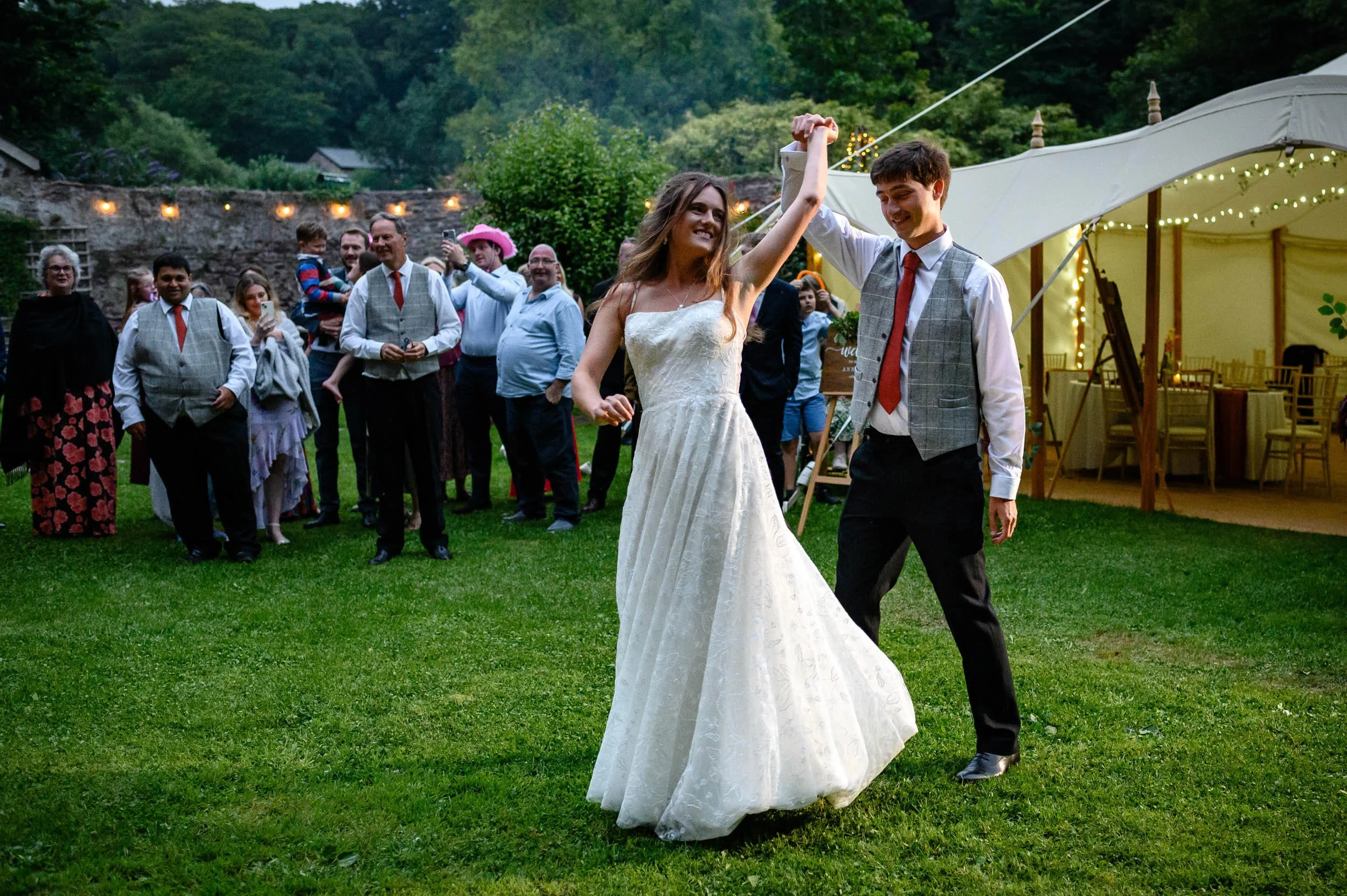 A bride and groom dancing at their walled garden wedding reception outdoors during the evening, surrounded by guests, with a decorated tent and trees in the background.