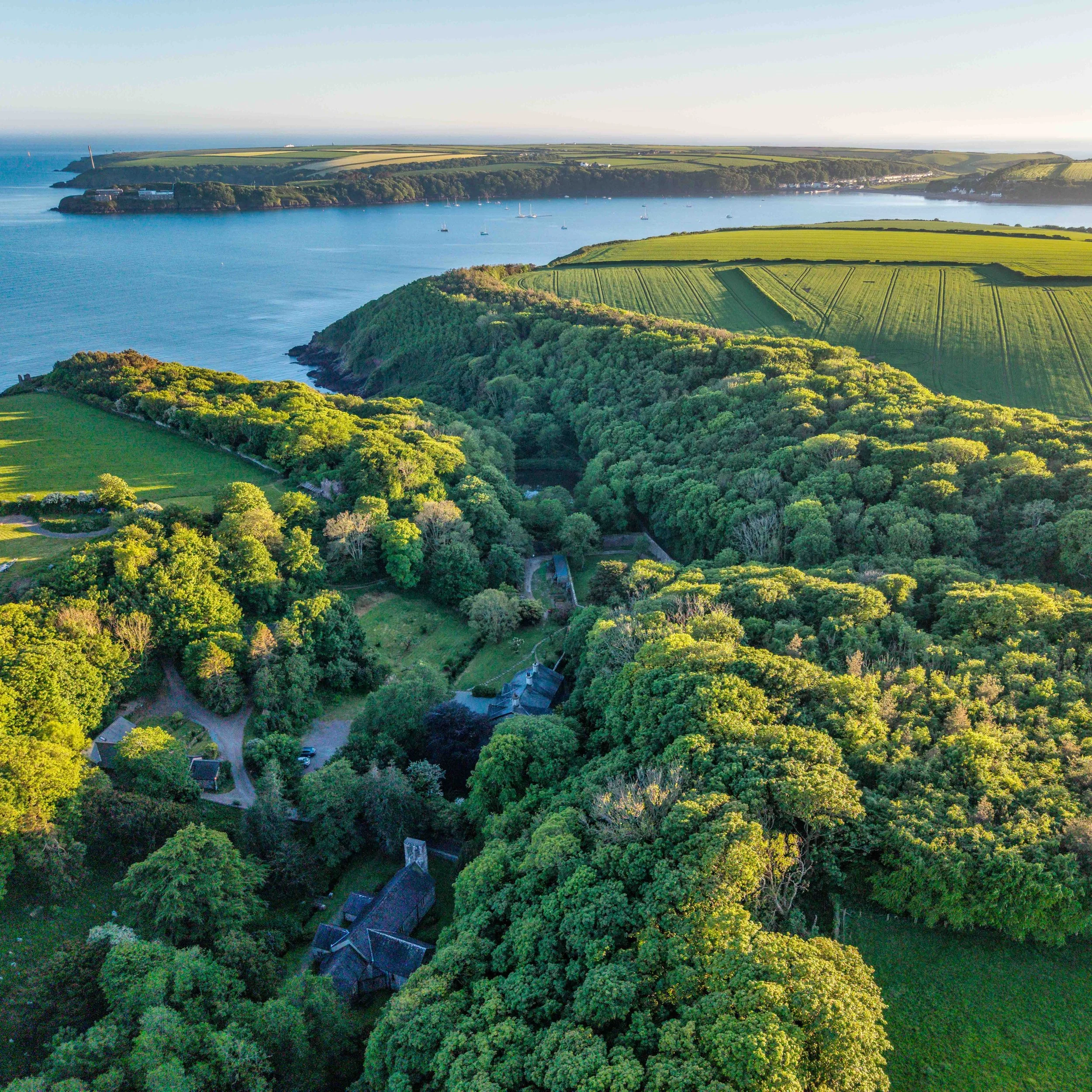 Aerial view of a coastal landscape with lush green trees, fields, and a body of water with sailboats, under a clear sky.