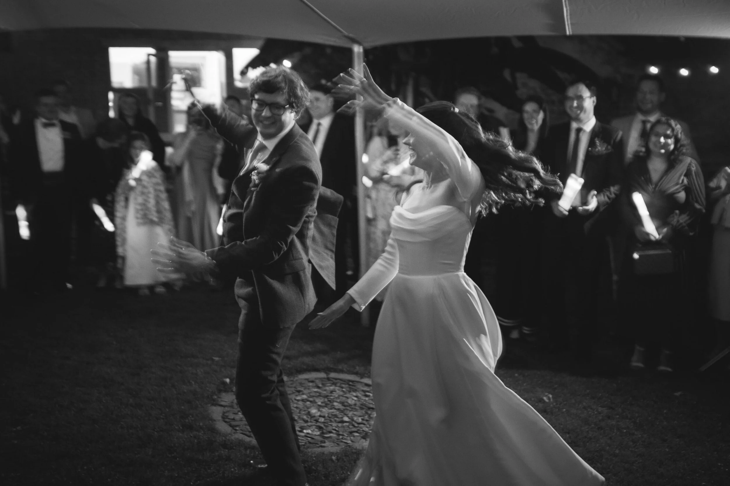 A black and white photo of a wedding reception with a bride and groom dancing happily, surrounded by guests.