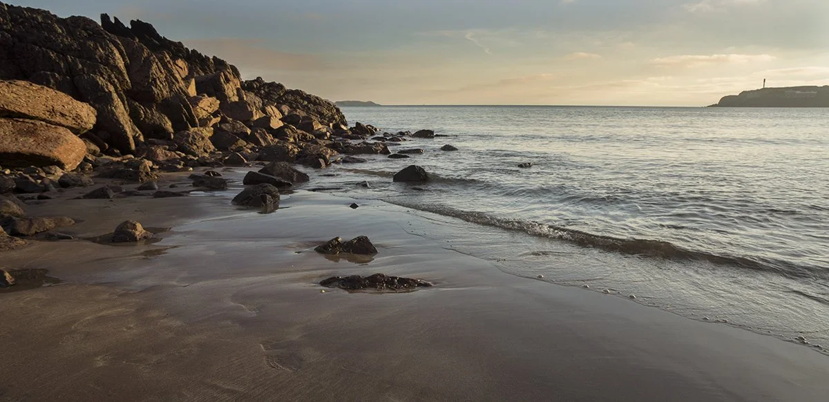 A rocky shoreline with waves gently lapping onto the sandy beach, with a distant landmass and a lighthouse in the background during sunset.