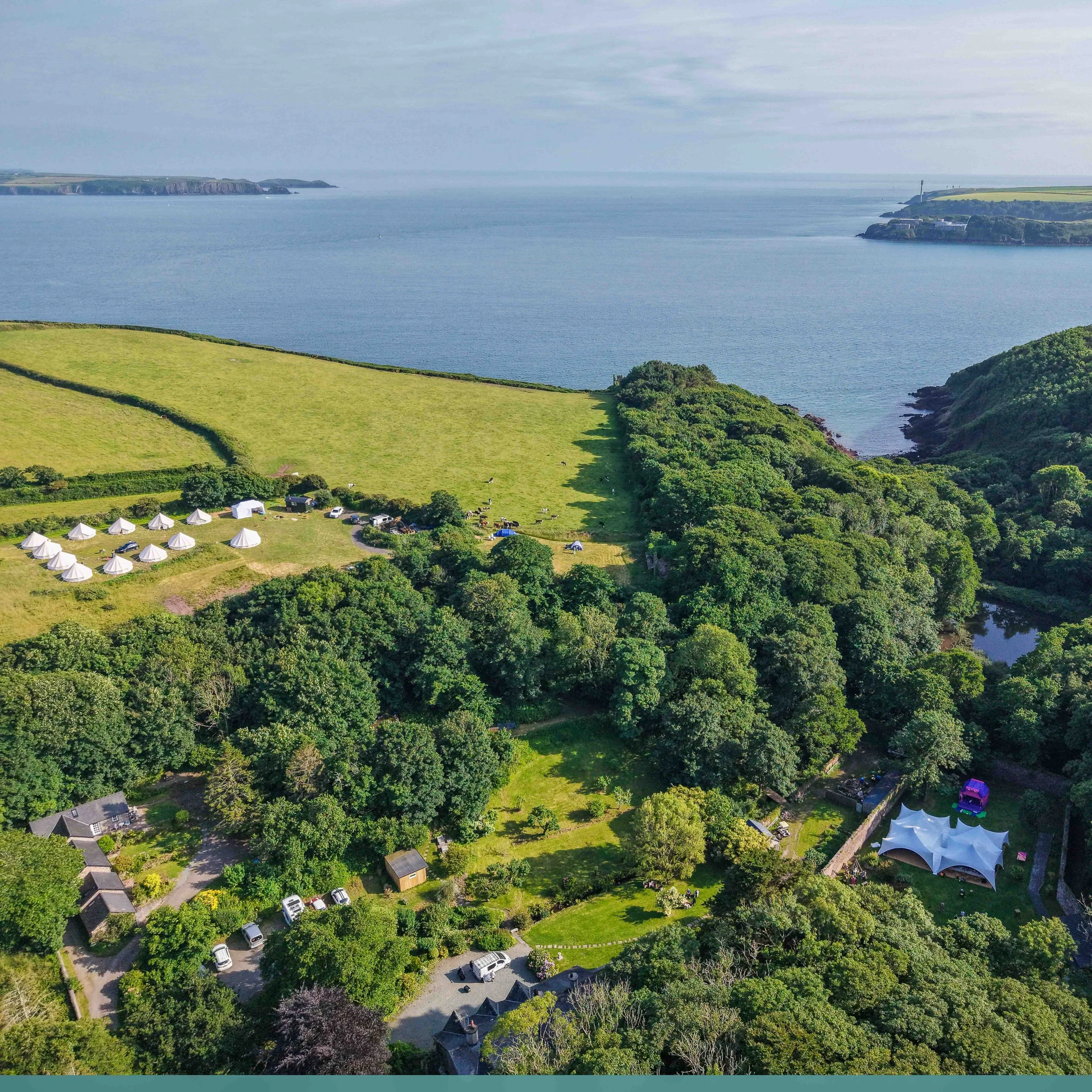 Aerial view of a coastal landscape with green fields, trees, houses, and tents near the shoreline and a large body of water with cliffs and landmasses in the background.