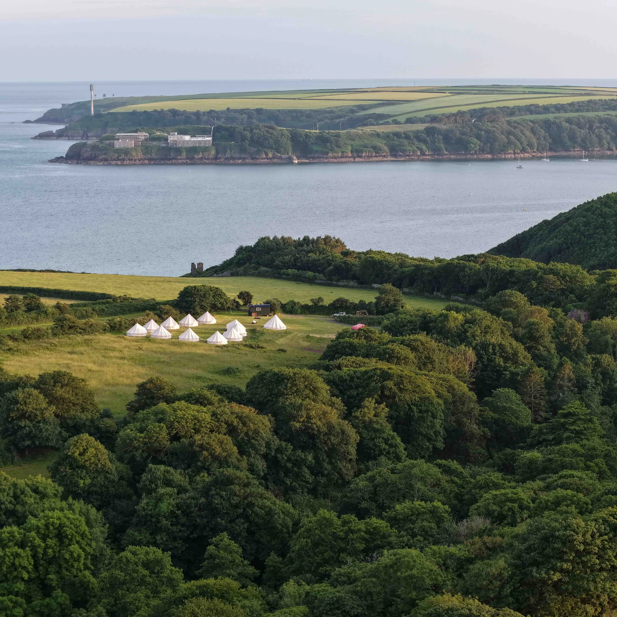 A scenic view of a coastal landscape with lush green trees, a body of water, and rolling fields extending towards the horizon. There are white tents set up on a grassy area in the foreground, and some small boats on the water.