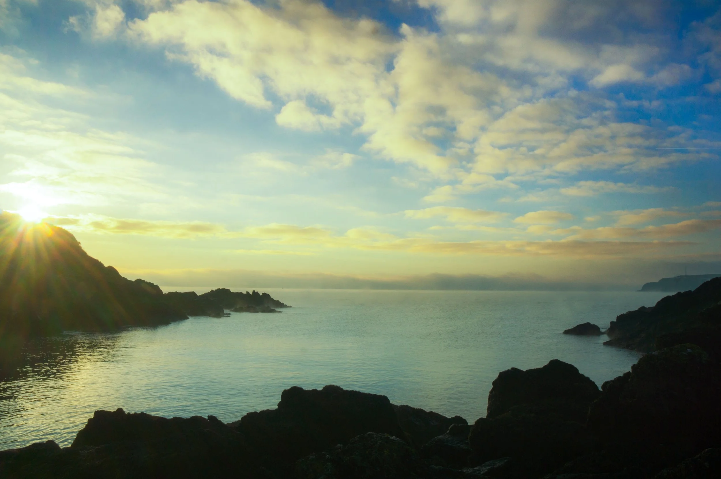 Sunrise over a coastal landscape with rocky cliffs, calm water, and a partly cloudy sky.