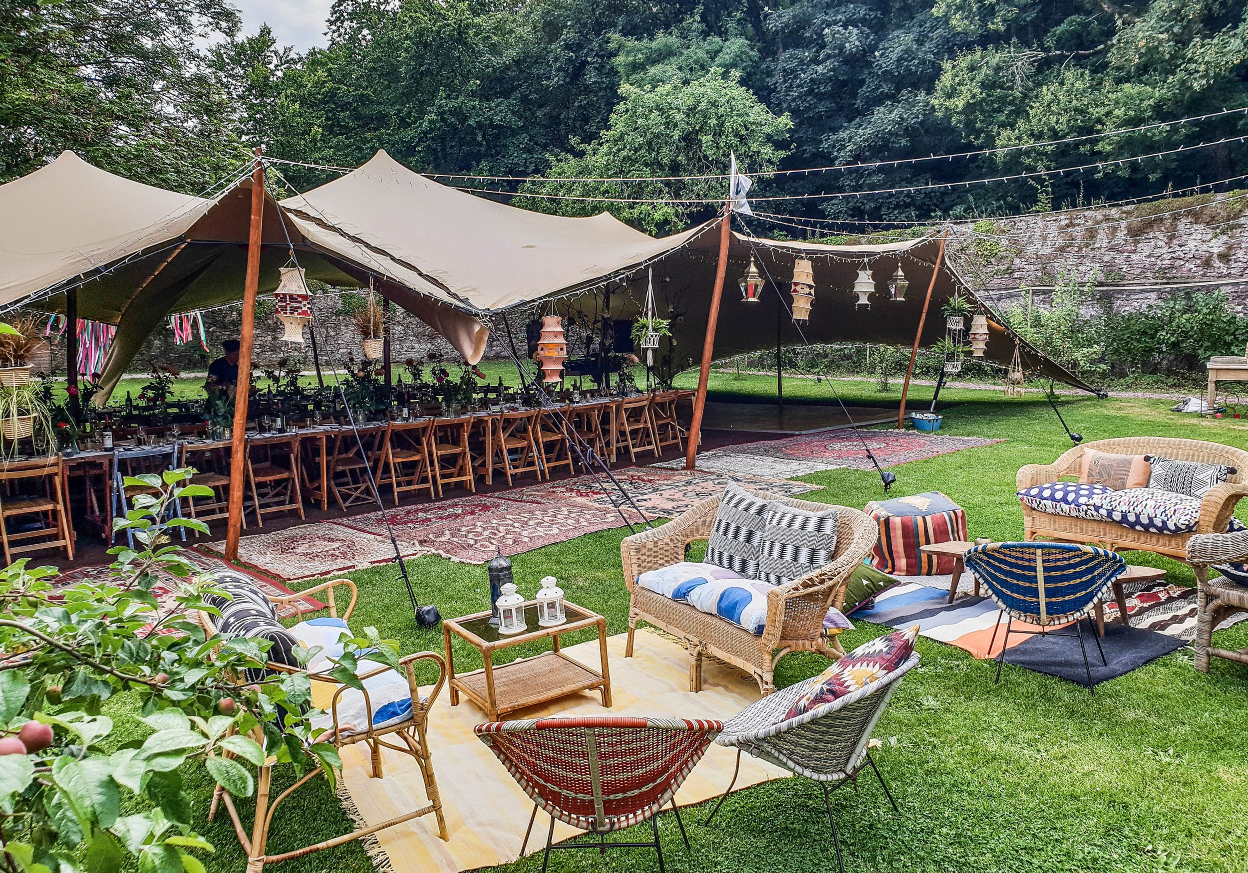 An outdoor garden party setup with a long table under a large beige canopy, surrounded by lounge chairs, rugs, and string lights, with trees and a stone wall in the background.