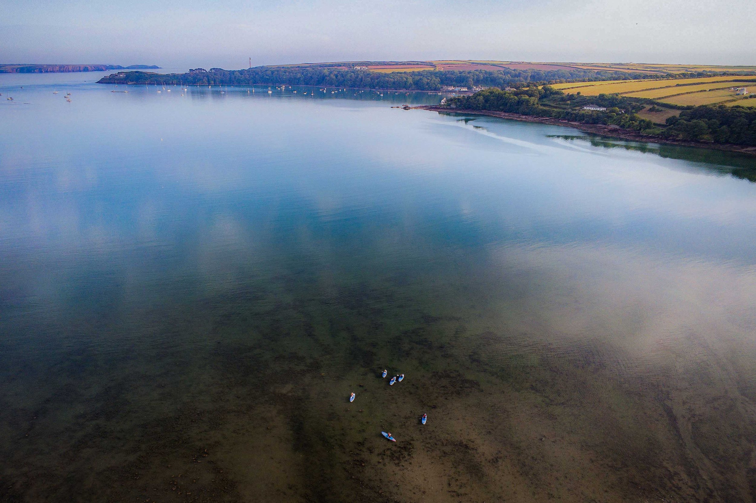 Aerial view of a calm blue bay with multiple small sailboats and kayaks floating on the water. The coastline features lush greenery and rolling hills with hints of yellow fields and scattered white buildings. The water is clear near the shore, reveal