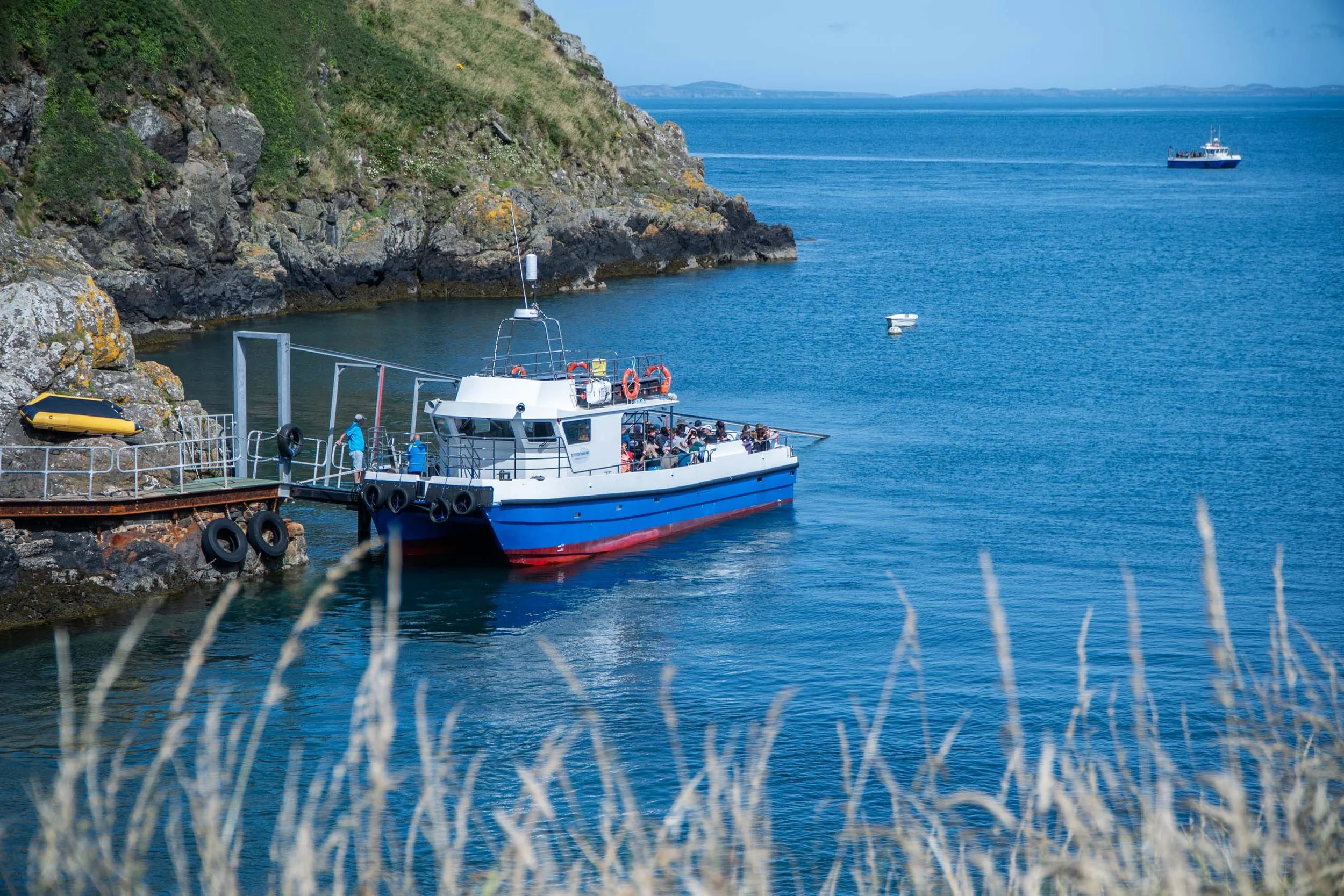 A small boat with passengers docked at a rocky shoreline, with a large boat sailing in the distance on calm blue waters and a clear sky.