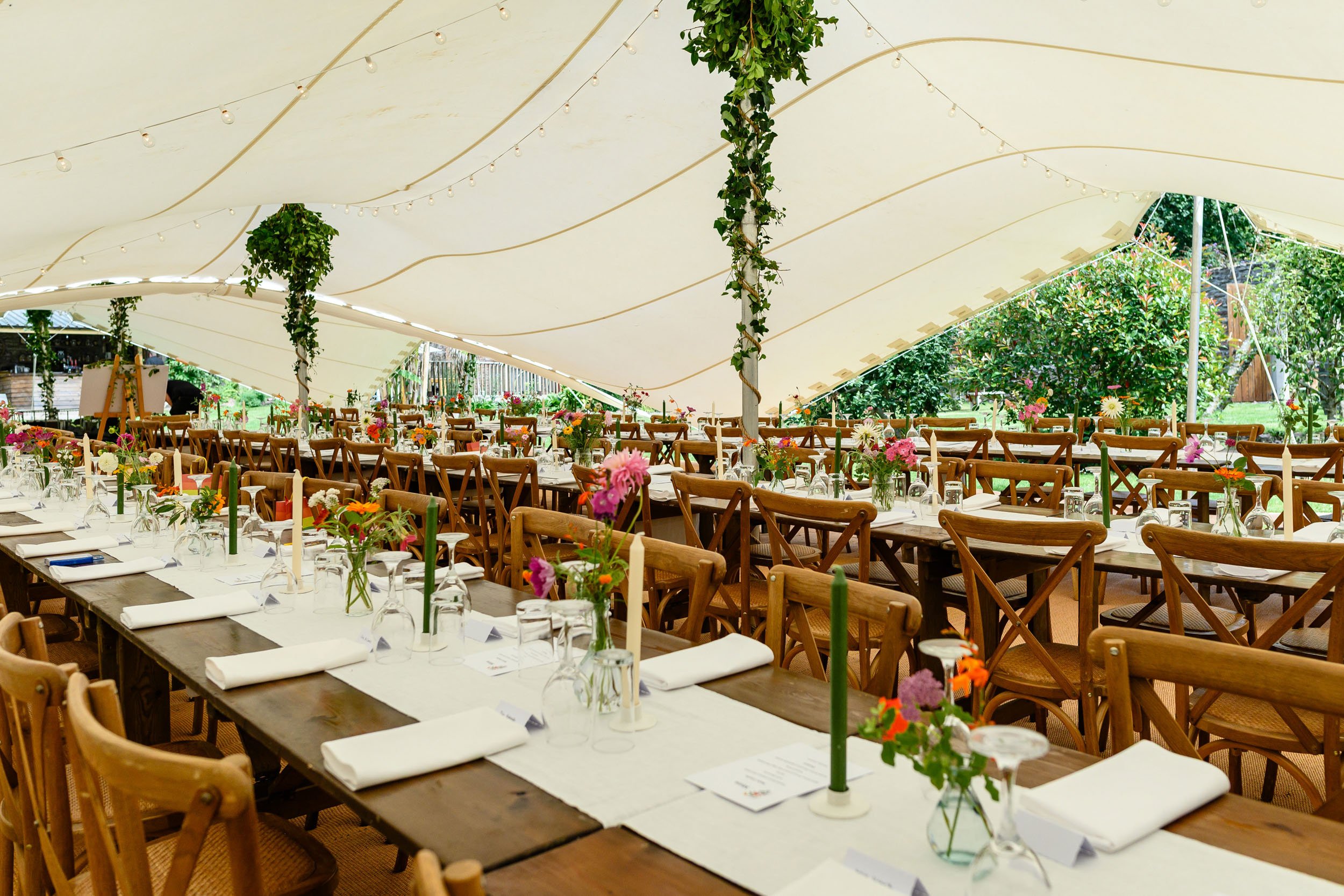 A large outdoor wedding reception area under a white tent with string lights, long tables decorated with flowers, and wooden chairs.