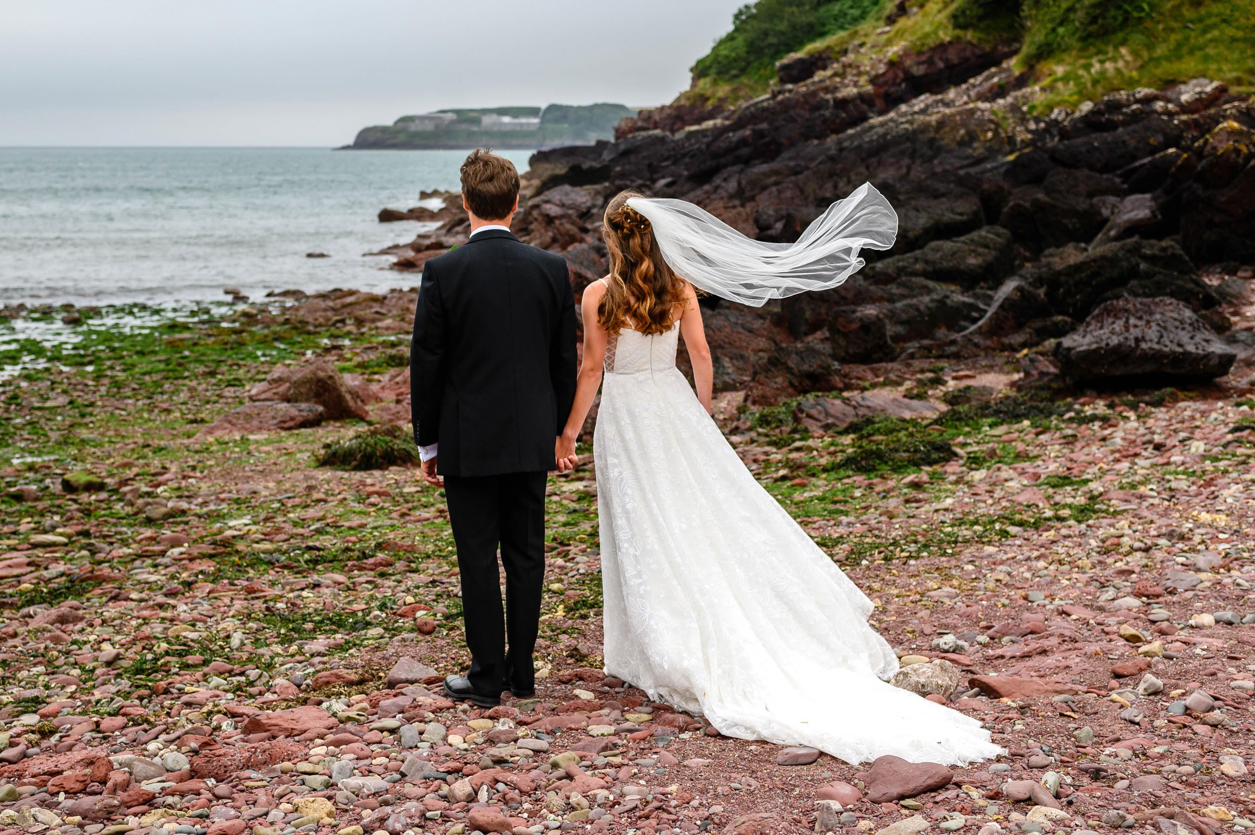 A bride and groom holding hands on a rocky beach, with the bride wearing a wedding dress and veil, and the groom in a black suit, facing the ocean with cliffs in the background.