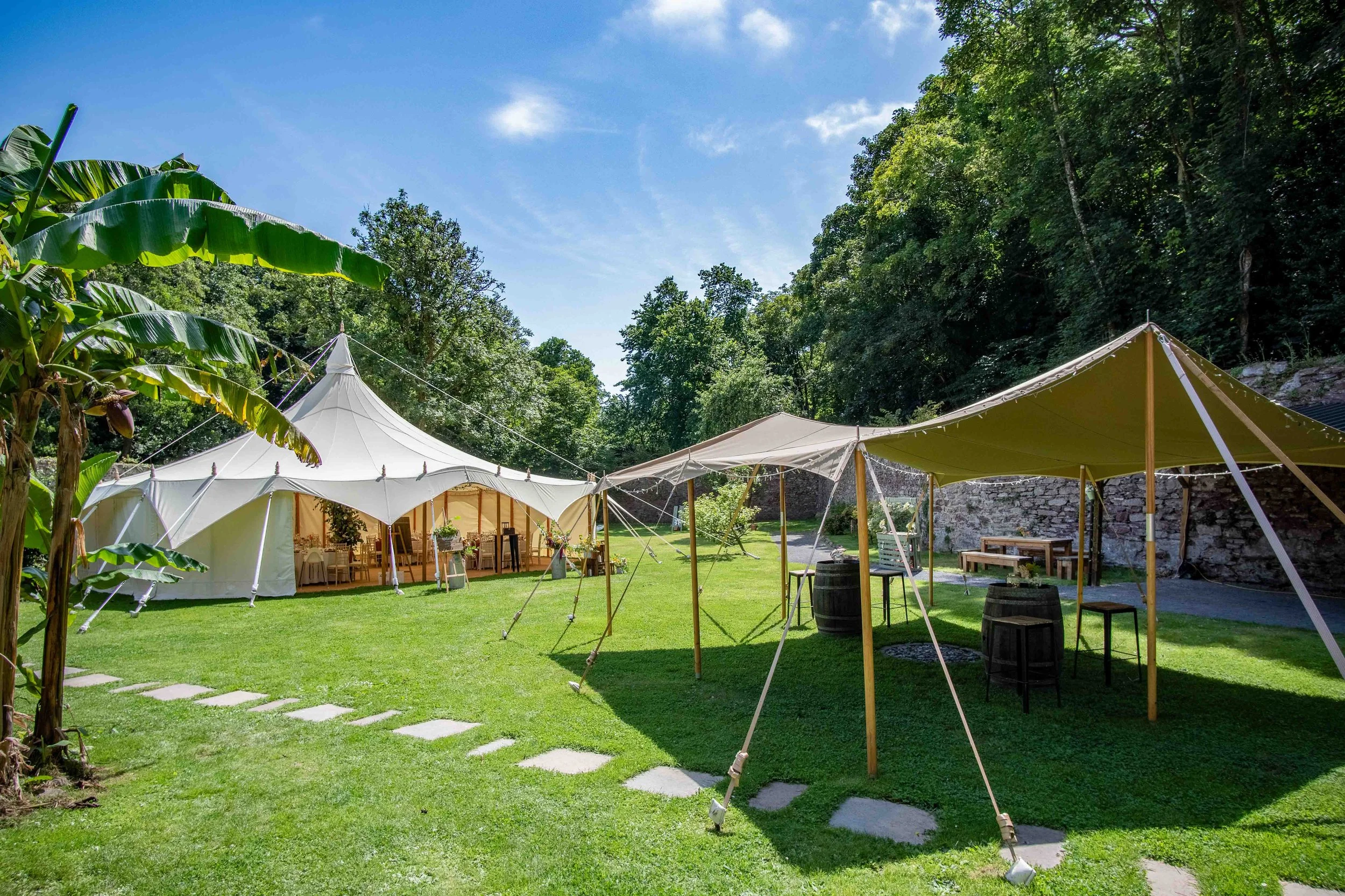 Outdoor event setup with white and beige canopy tents, tables, chairs, and string lights on a grassy area surrounded by trees under a blue sky.