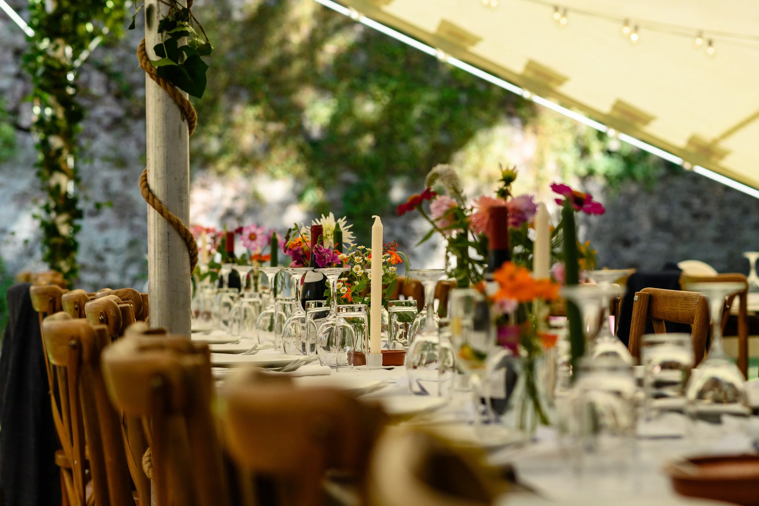 A decorated outdoor dining table with flowers, candles, and glasses, set under a canopy with string lights, surrounded by wooden chairs and greenery.