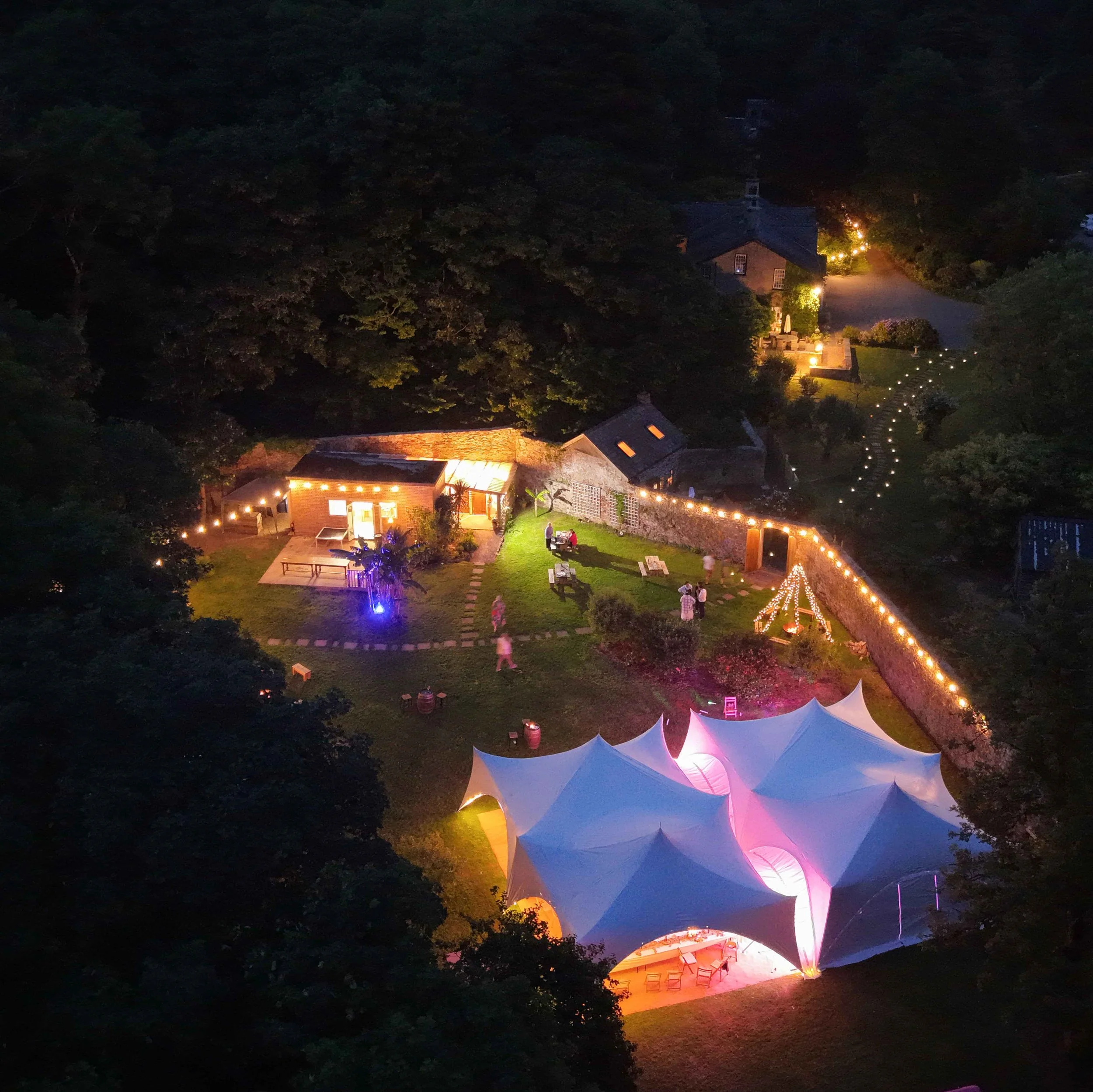 Night scene featuring a house illuminated with warm lights, surrounded by trees and a string of decorative lights forming a pathway, with a tent-like structure in the foreground and a few people visible. The area appears to be part of an outdoor even