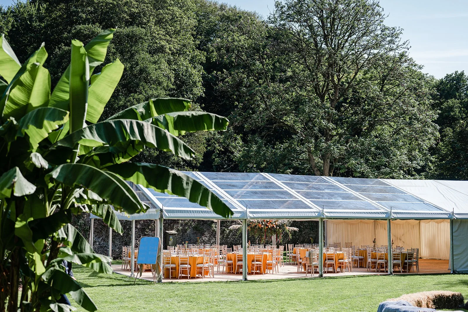 A large outdoor tent with chairs and tables inside, set up for an event, surrounded by green trees and plants.
