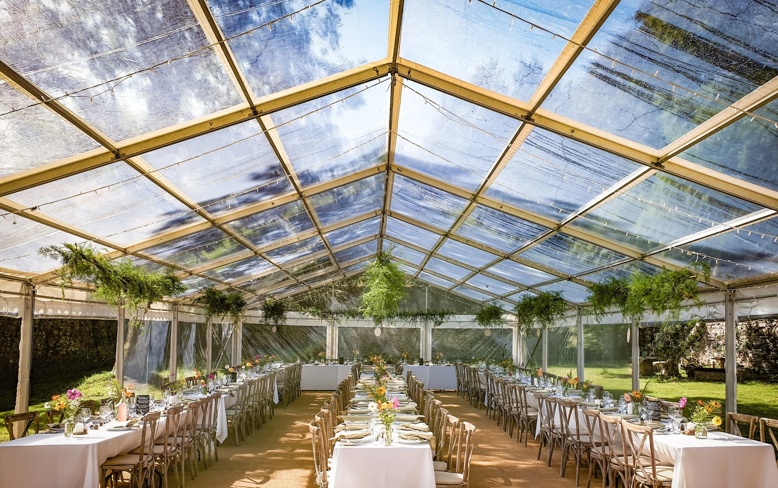 Long banquet tables inside a clear marquee, decorated with flowers, set for a meal with chairs around them. The tent has a transparent roof revealing the sky, and there are green plants hanging inside.