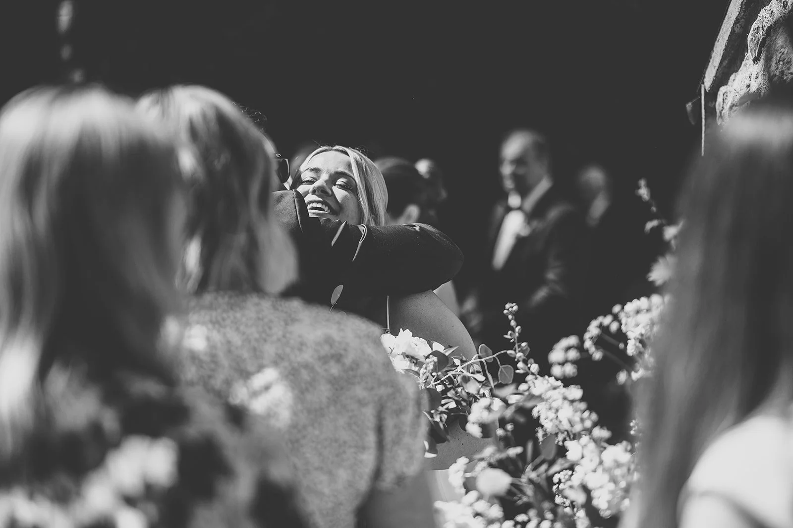 Black and white photo of a woman smiling and hugging another person at a wedding, surrounded by guests.